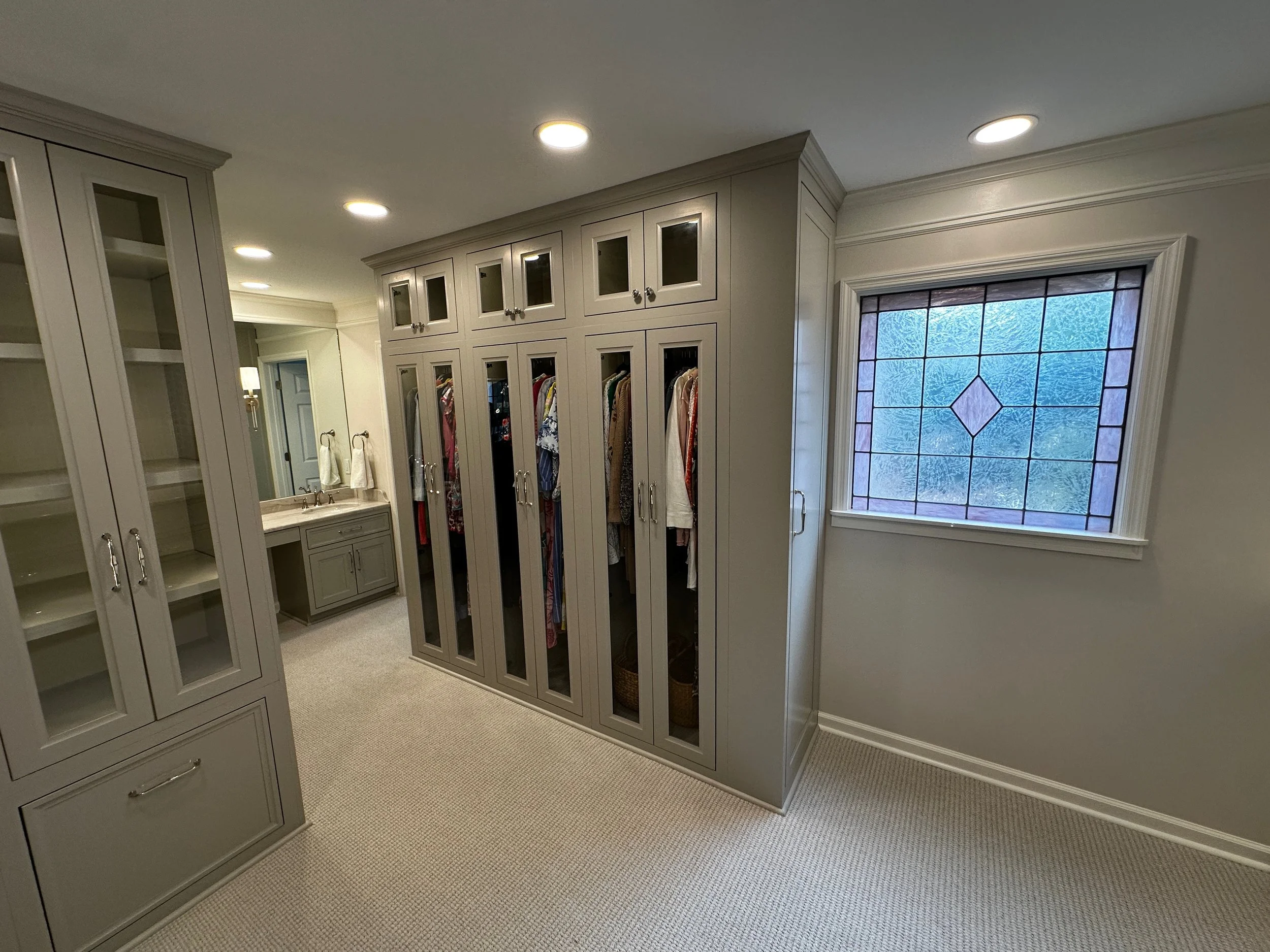 A spacious walk-in closet with built-in cabinets and a window with stained glass. The closet contains hanging clothes behind glass doors and a small vanity with a mirror and towel hooks.