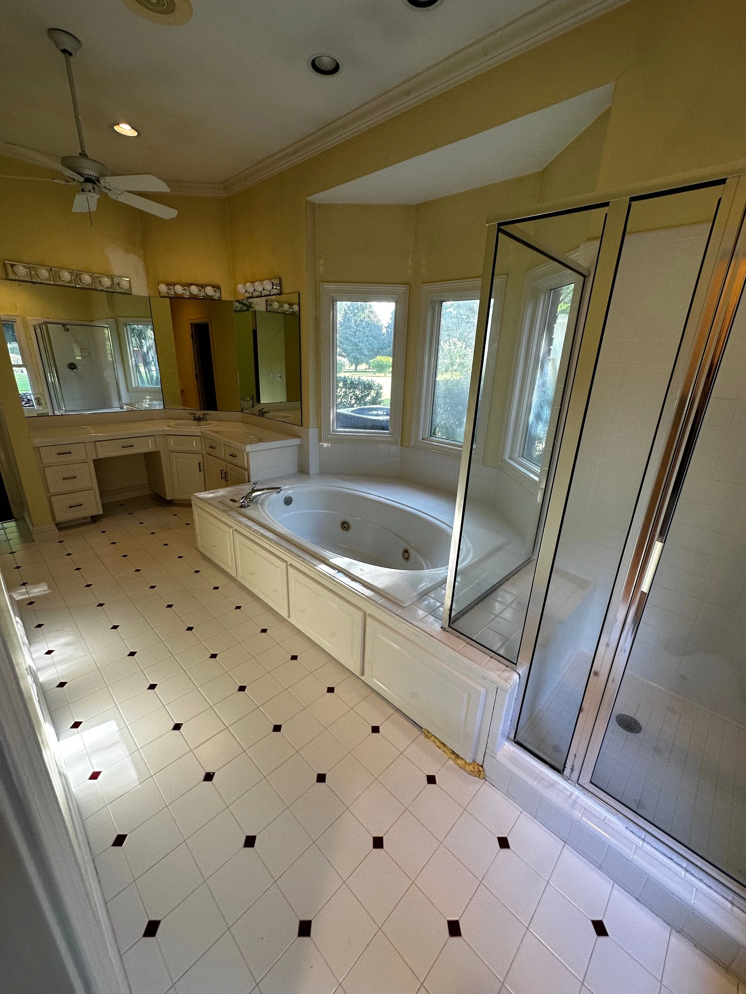Bathroom with a large corner bathtub and a separate glass-enclosed shower, vanity mirrors with lights, and windows showing a green outdoor view.