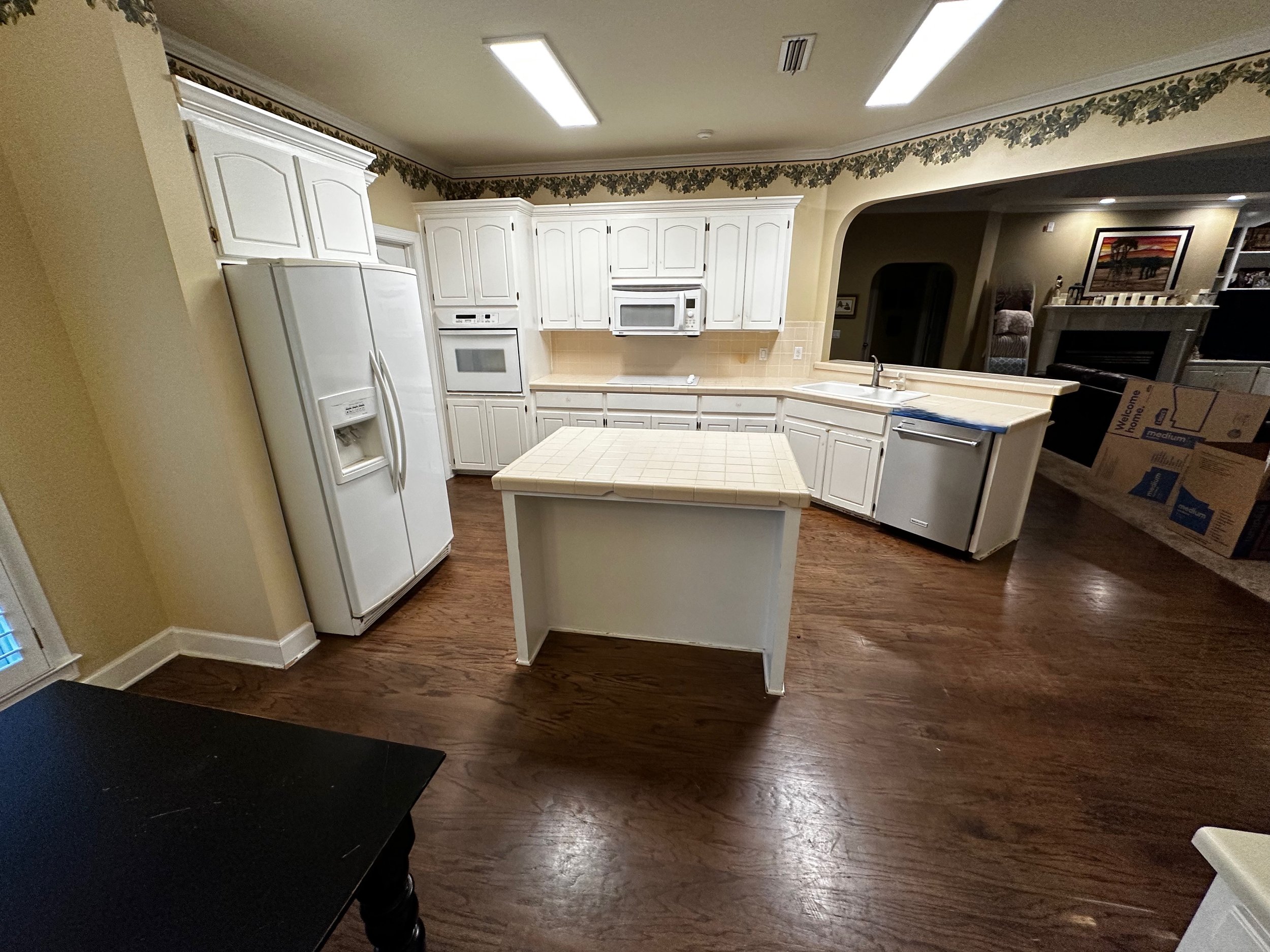 Empty kitchen with white cabinets, refrigerator, microwave, dishwasher, and a small island in the center. Wooden floor and a view into the living room with boxes and furniture.