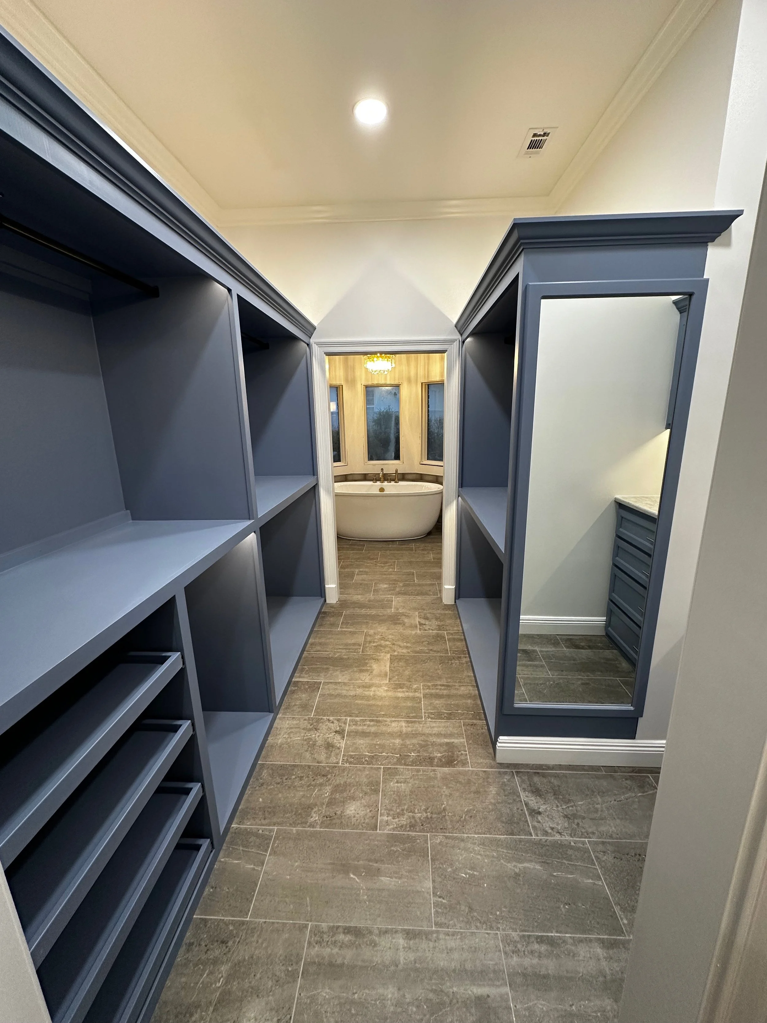 Empty closet with blue shelving on the left, a large mirror on the right wall, and a bathroom with a bathtub at the end of the hallway. The floor has brown tile, and the ceiling has a light and an air vent.
