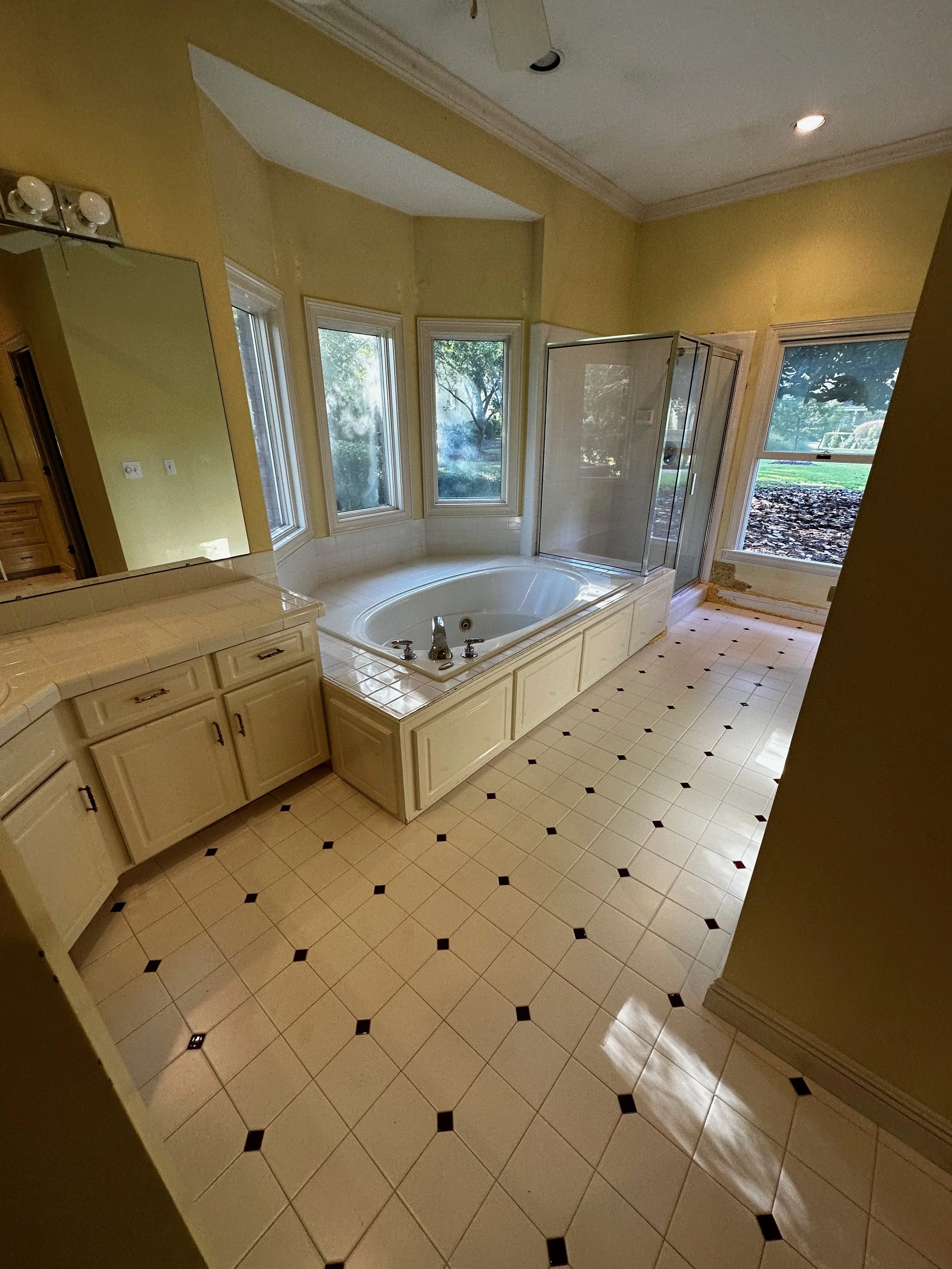 A spacious bathroom with a large bathtub under three windows, a built-in vanity with a mirror, a glass shower stall, and a floor with a white tile pattern featuring small black accents.
