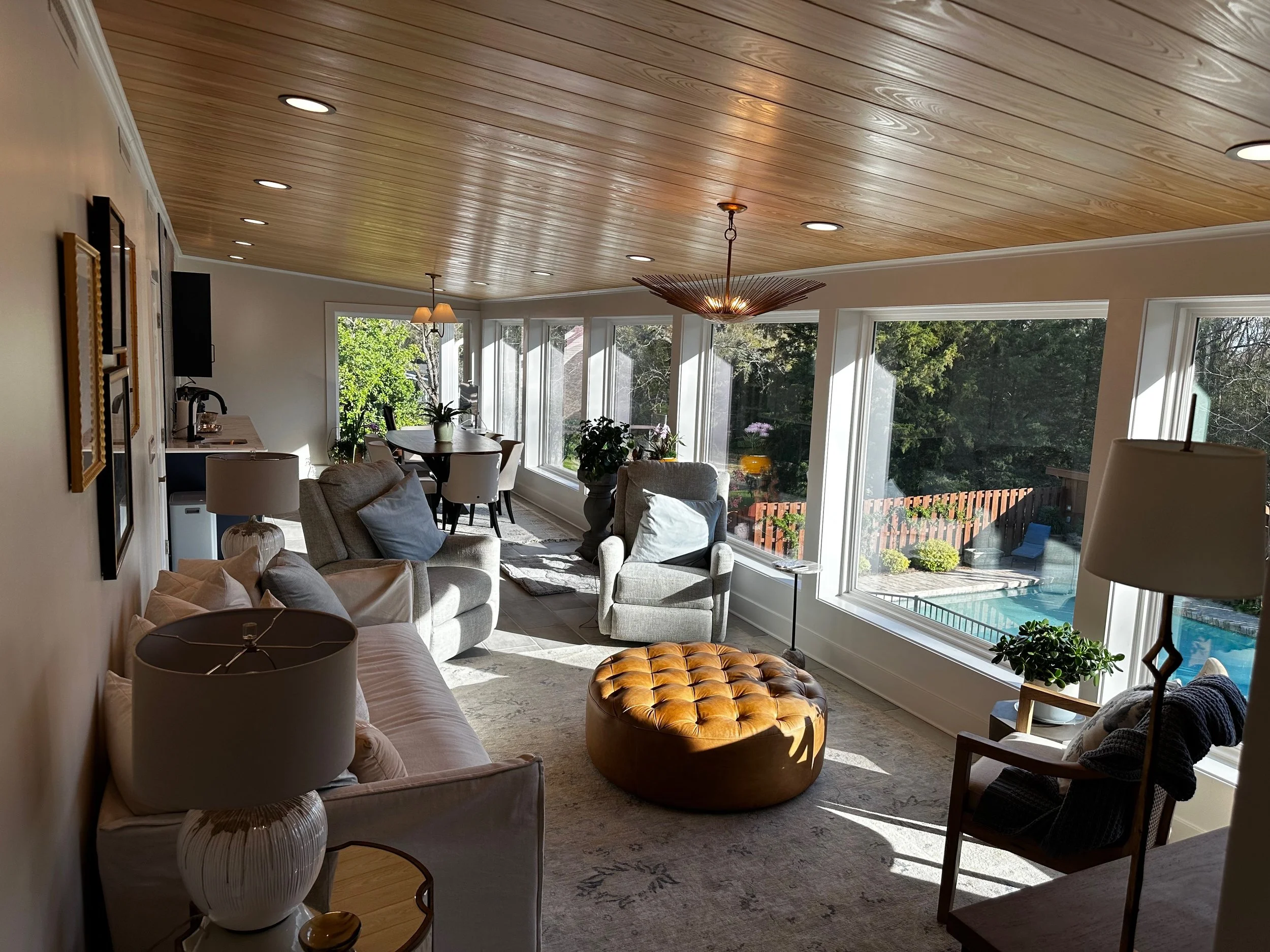 Sunlit living room with large windows, gray armchairs, a tufted leather ottoman, a sofa, a dining table with chairs, and a view of a backyard with a pool.