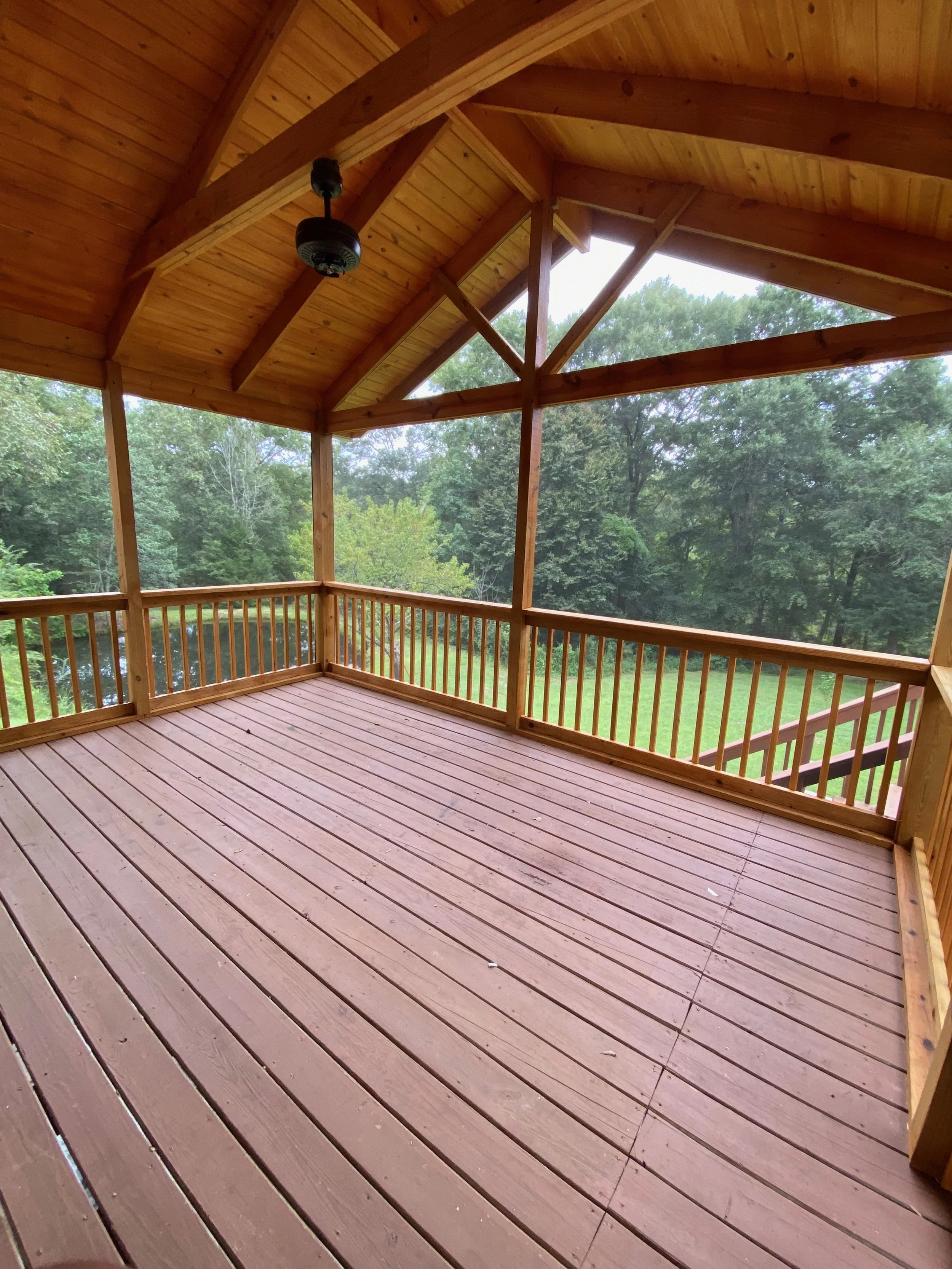 Image of a wooden screened-in porch with a ceiling fan, overlooking a lush green wooded area.