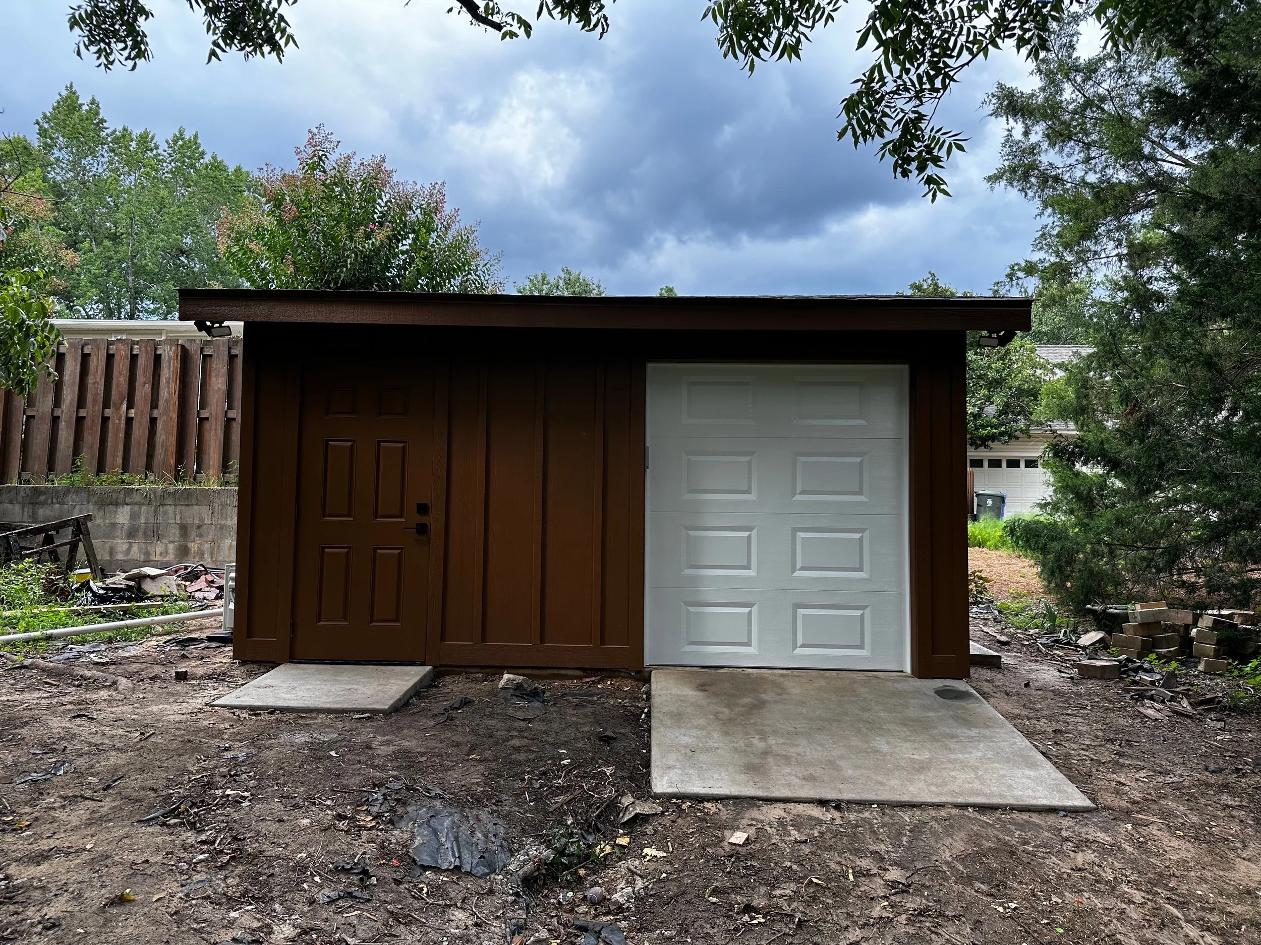 A small brown building with a single brown door and a white garage door, situated on a dirt ground with concrete ramps in front of each door, surrounded by trees and a cloudy sky.