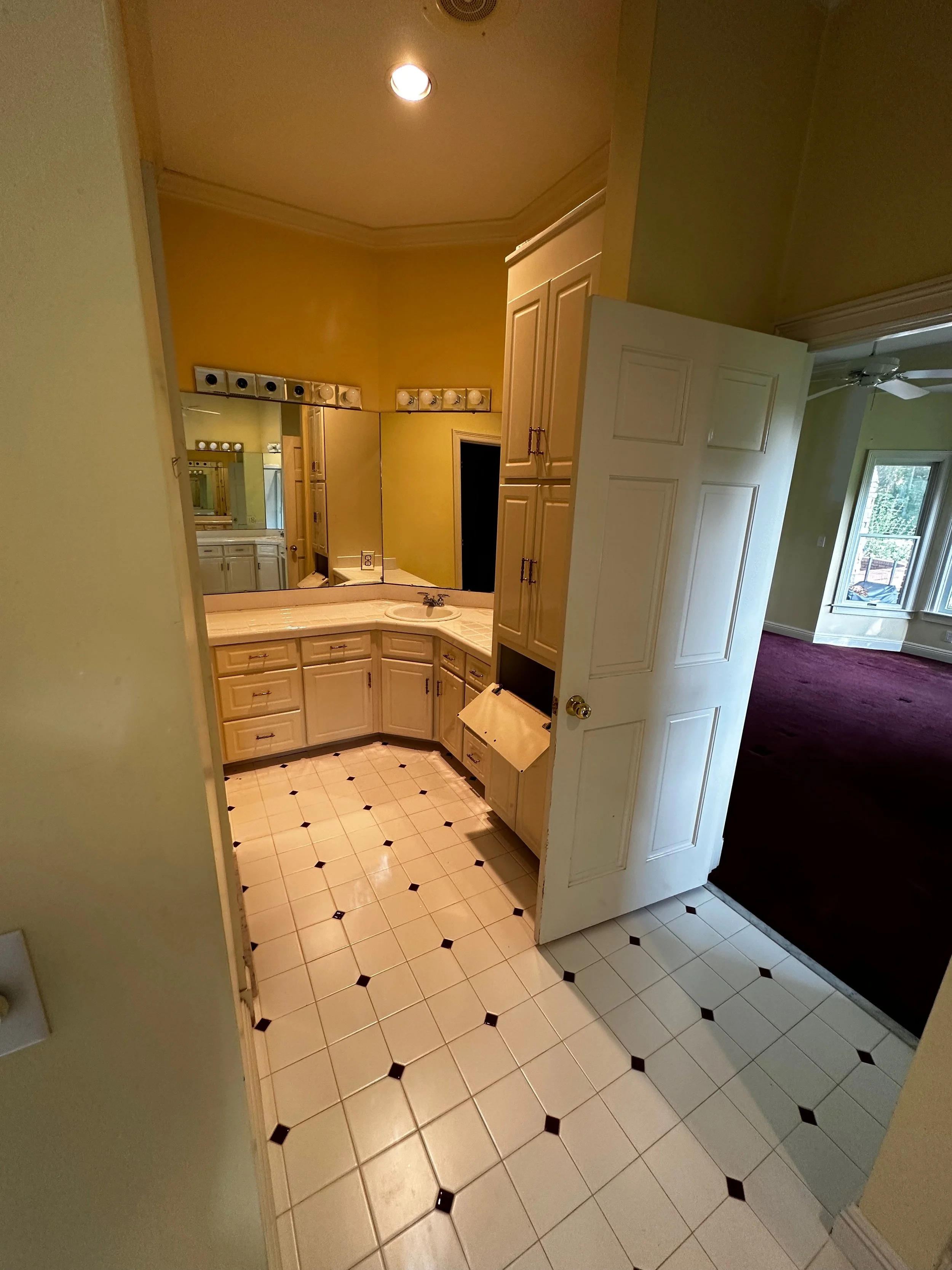 Bathroom with white cabinetry, a mirror with light bulbs, and a beige curved countertop. The door to the bathroom is partially open, revealing an adjacent room with a purple carpet and large windows.