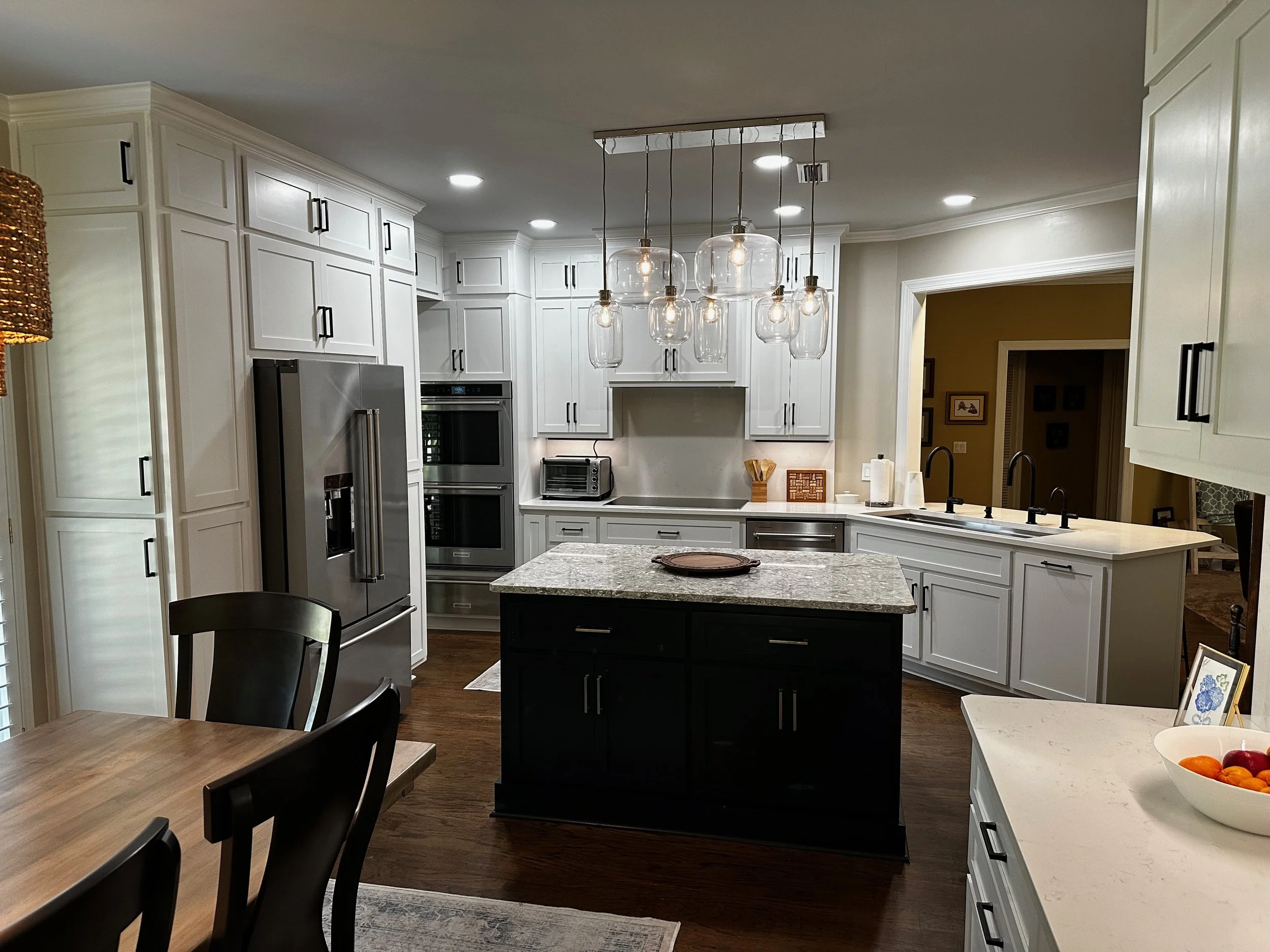 Modern kitchen with white cabinets, stainless steel refrigerator, black kitchen island with granite countertop, hanging pendant lights, and a dining table with black chairs.