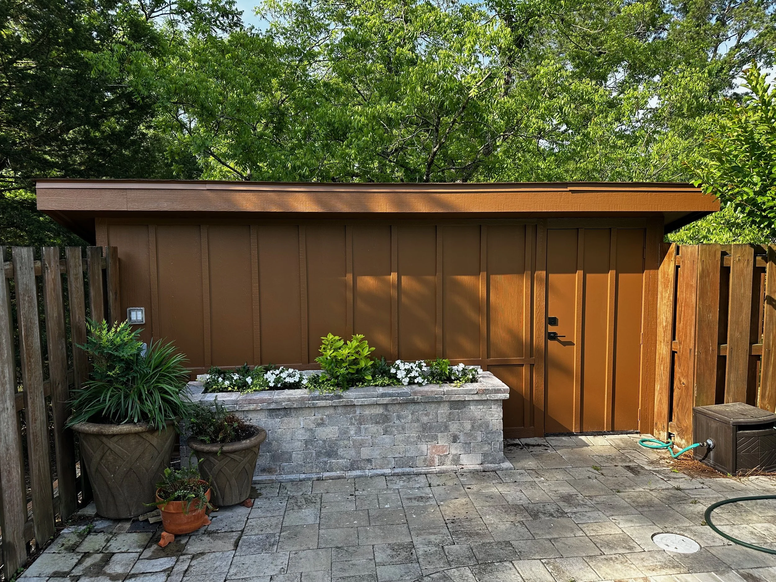 A small backyard shed painted brown, with a black door handle, surrounded by potted plants and a flower bed with white flowers. There is a brick planter box in front of the shed, and the backyard is paved with stone tiles. A garden hose and an outdoo