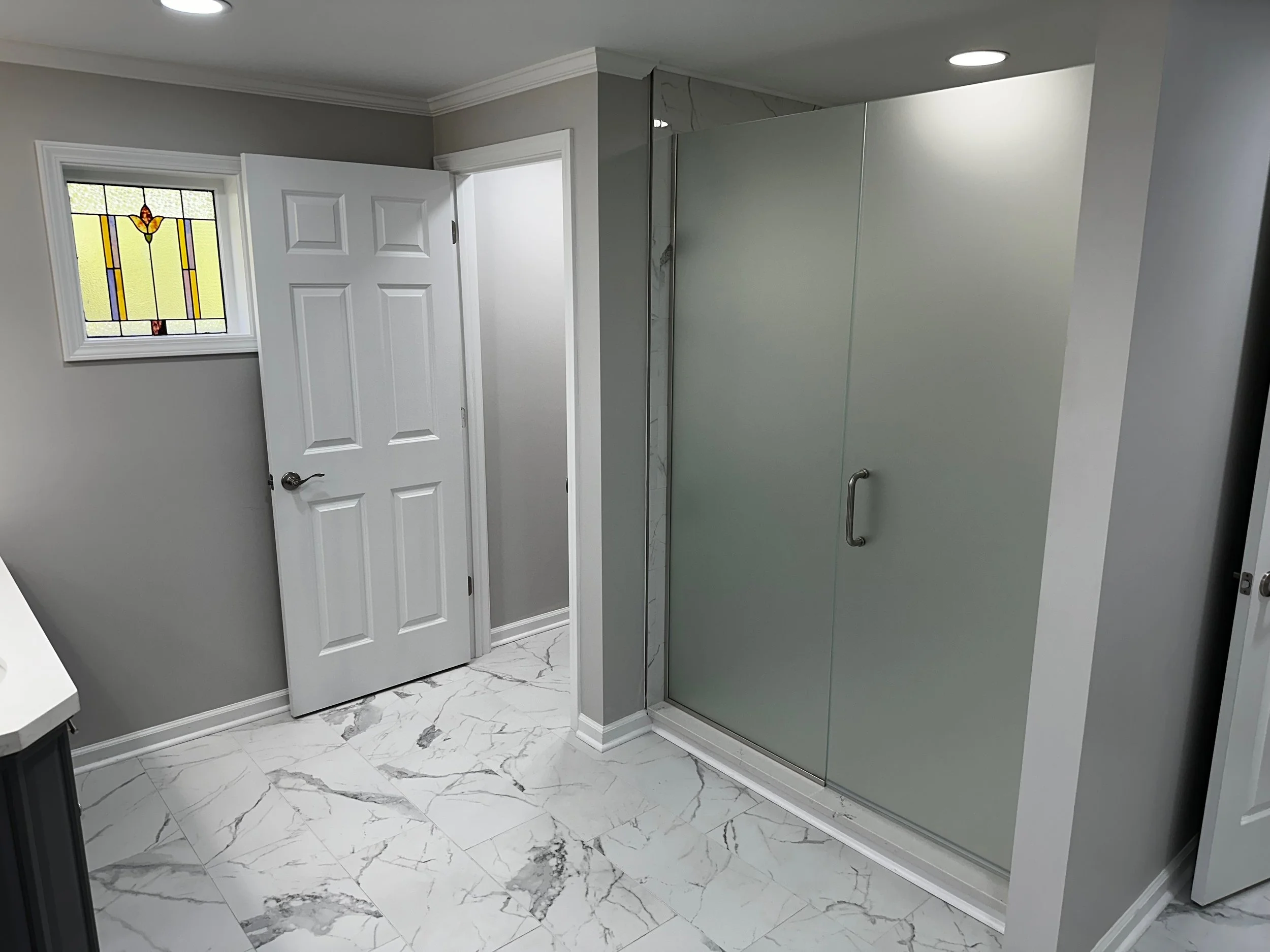 Modern bathroom with gray walls, a small stained glass window, a closed white door, and a frosted glass shower enclosure with a metal handle.