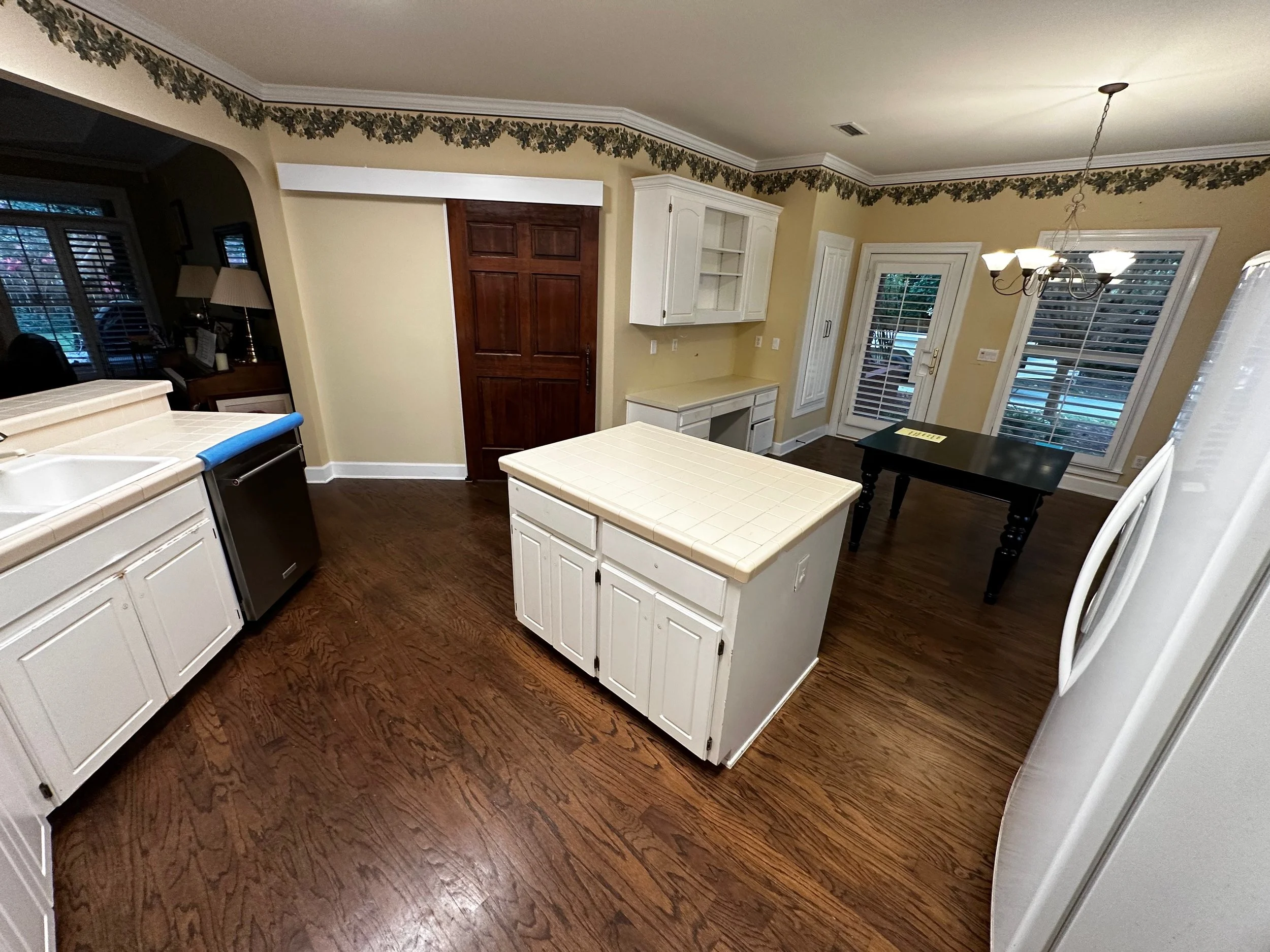 Empty kitchen with white cabinets, a small white island, a black dining table, hardwood floors, and a glass door leading outside.