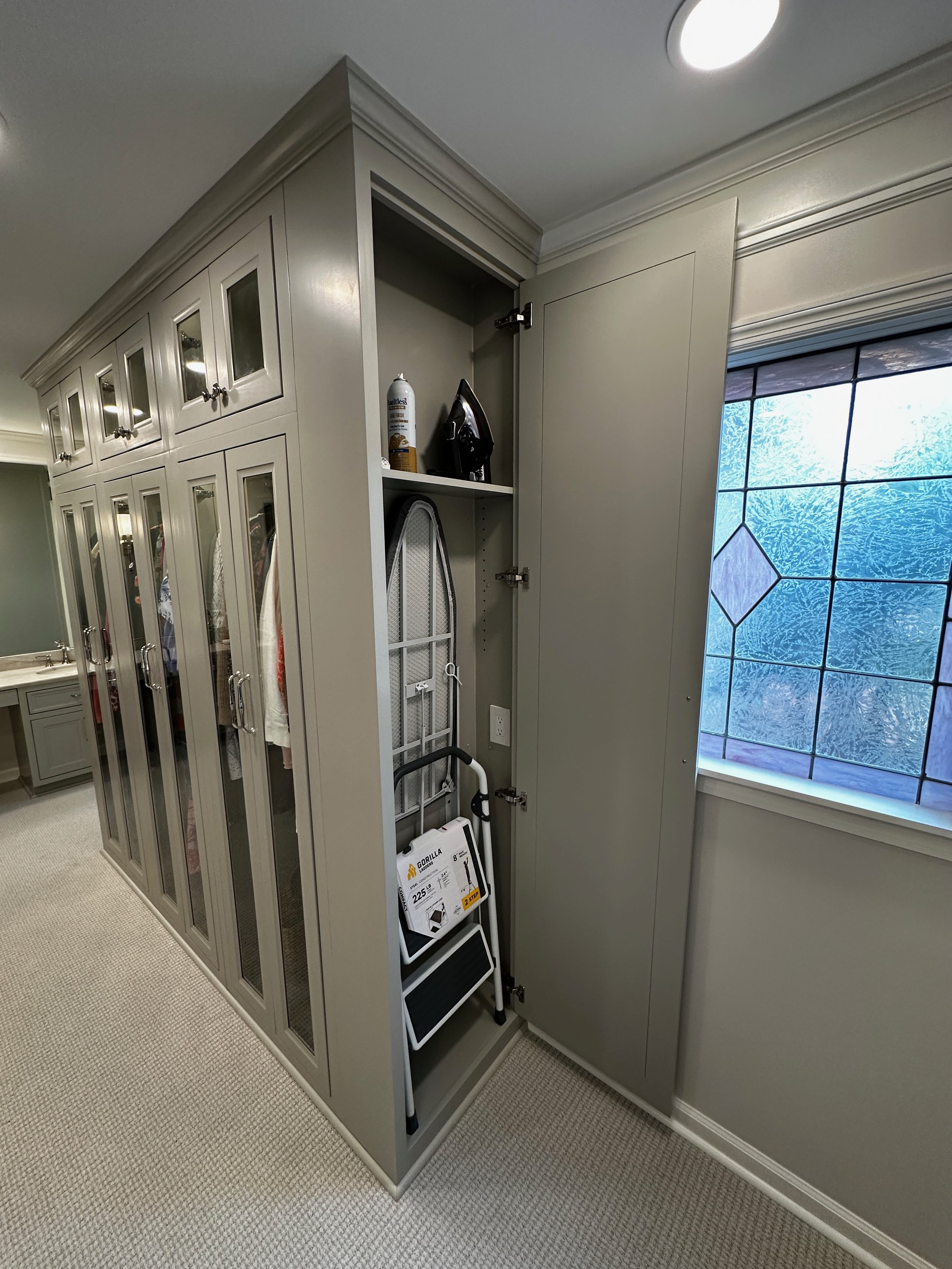 Closet with a step ladder and an iron, located near a window, in a room with a carpeted floor.