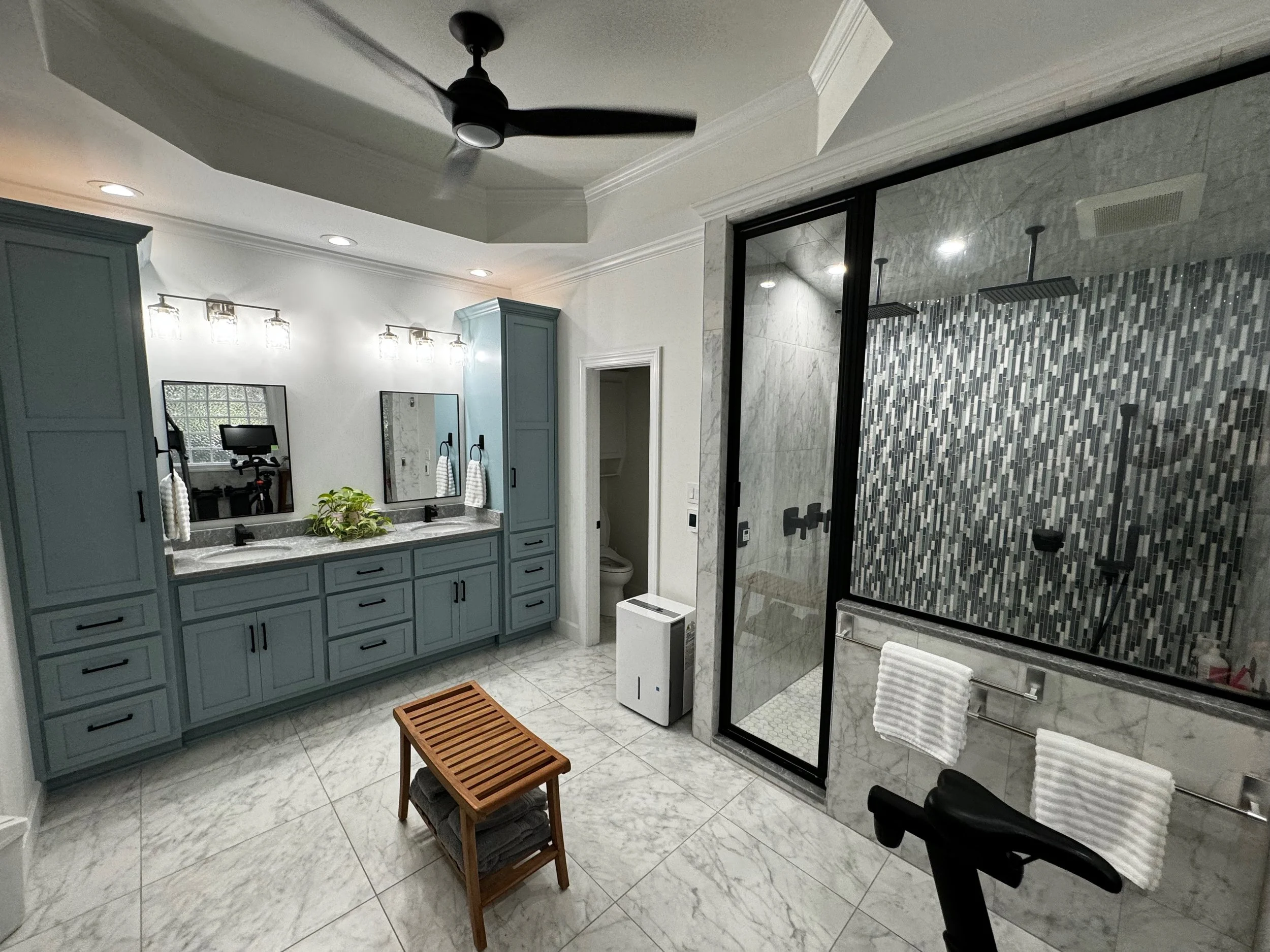 Modern bathroom with double vanity, blue cabinets, marble countertops, large mirrors, gray tiled walk-in shower with black fixtures, glass door, towel rack, and ceiling fan.