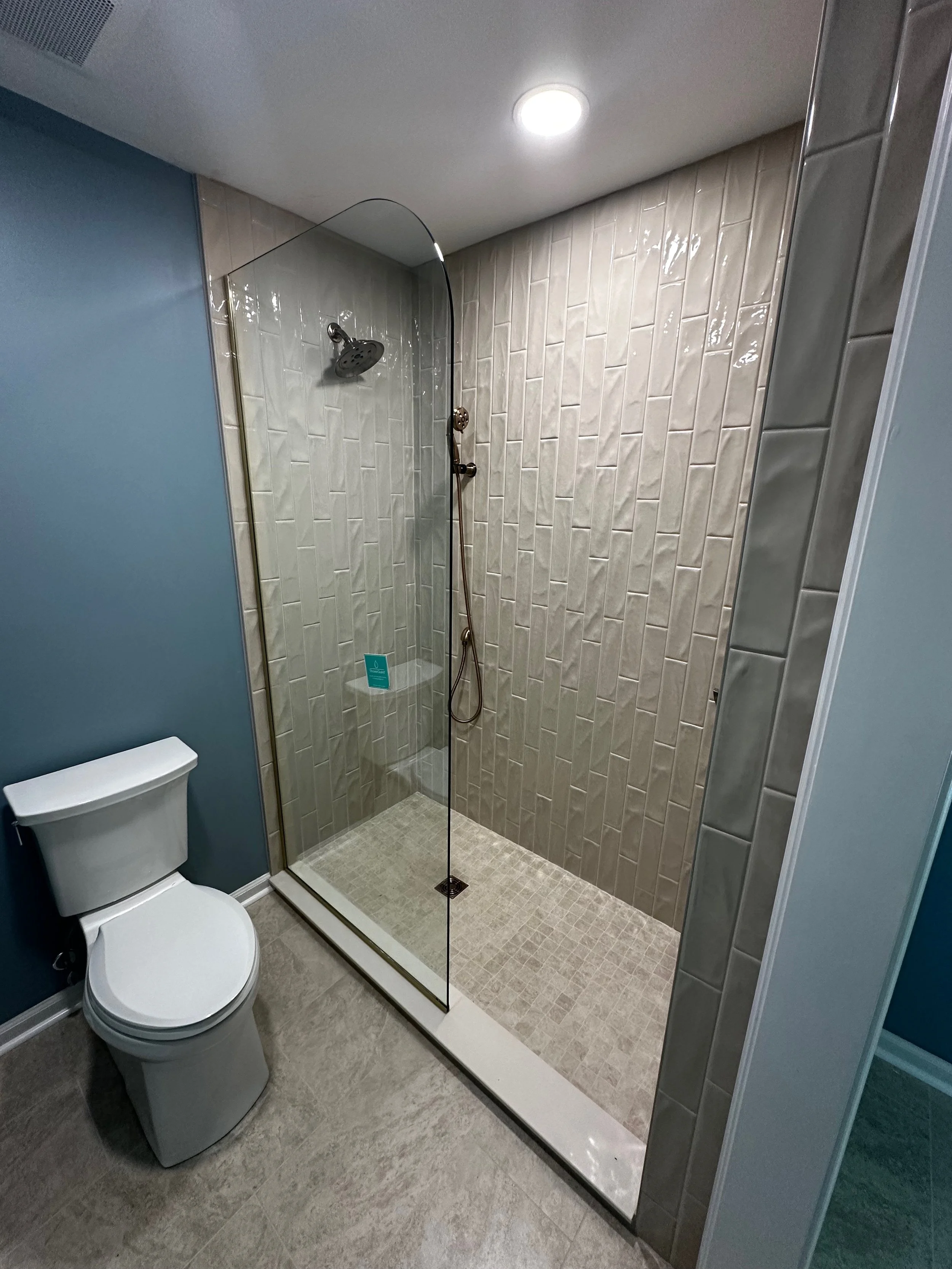 A bathroom with a glass shower enclosure, beige tiled walls and floor, a white toilet, and a ceiling light.