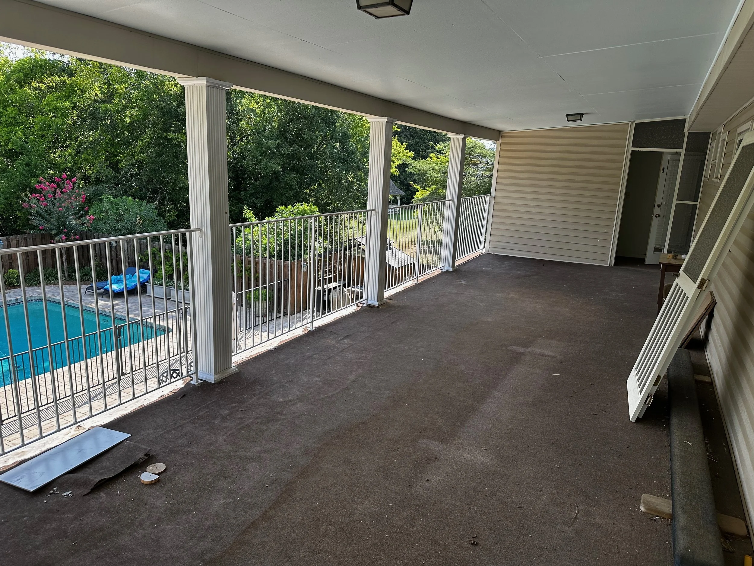 Empty outdoor balcony with brown carpet, white railing, beige siding, and green trees in the background, with a glimpse of a swimming pool and patio area below.