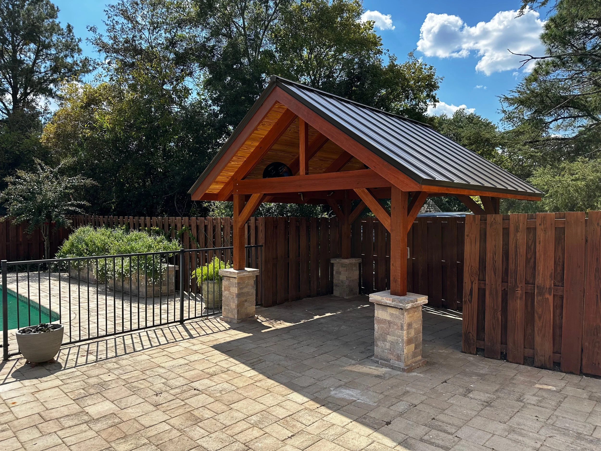 Photo of a backyard with a swimming pool, fenced with black railings, and a wooden picnic shelter with a metal roof and brick columns.