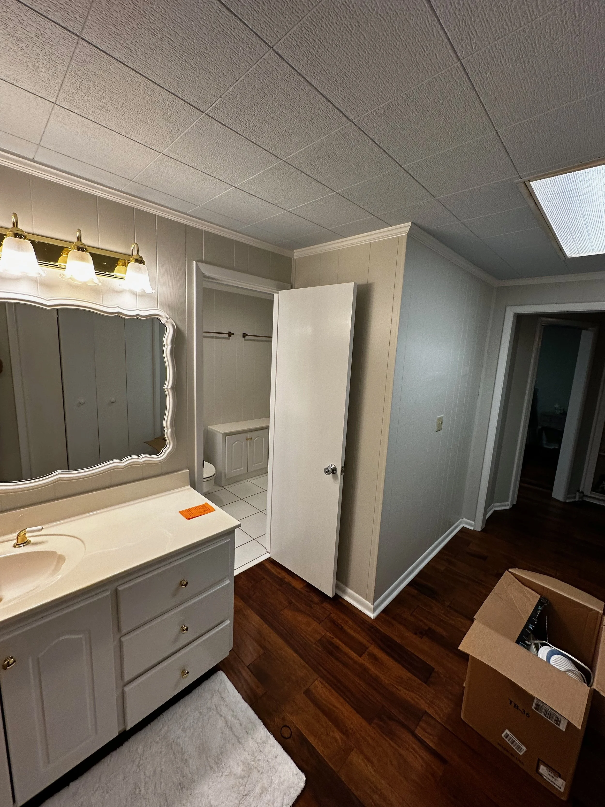 A bathroom with a cream-colored vanity with three drawers and a large mirror above it. A light fixture with three bulbs is mounted above the mirror. The bathroom has a white tiled floor and an open door revealing a separate room with a built-in cabin
