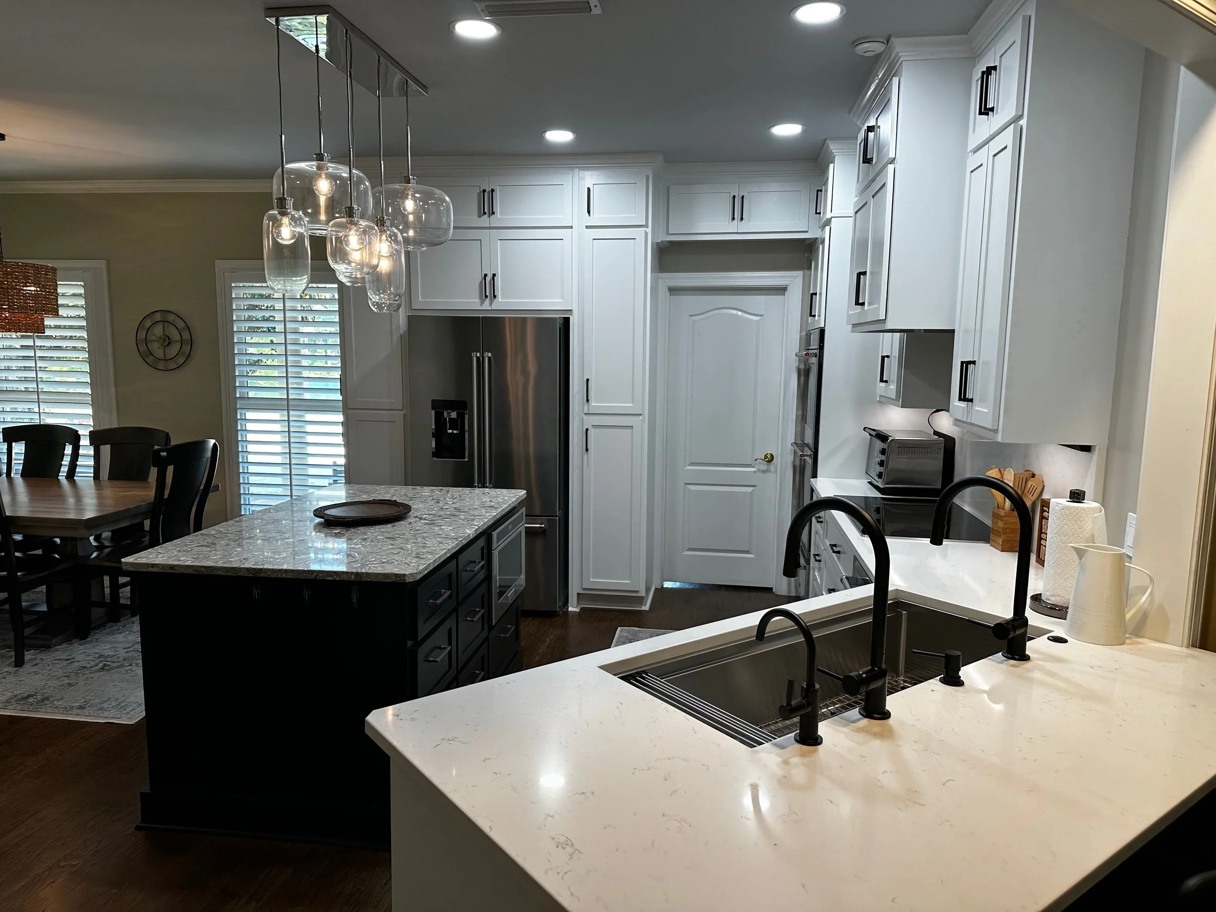 Modern kitchen with white cabinetry, black hardware, stainless steel appliances, a marble island, a coffee maker, and pendant lights over the island.