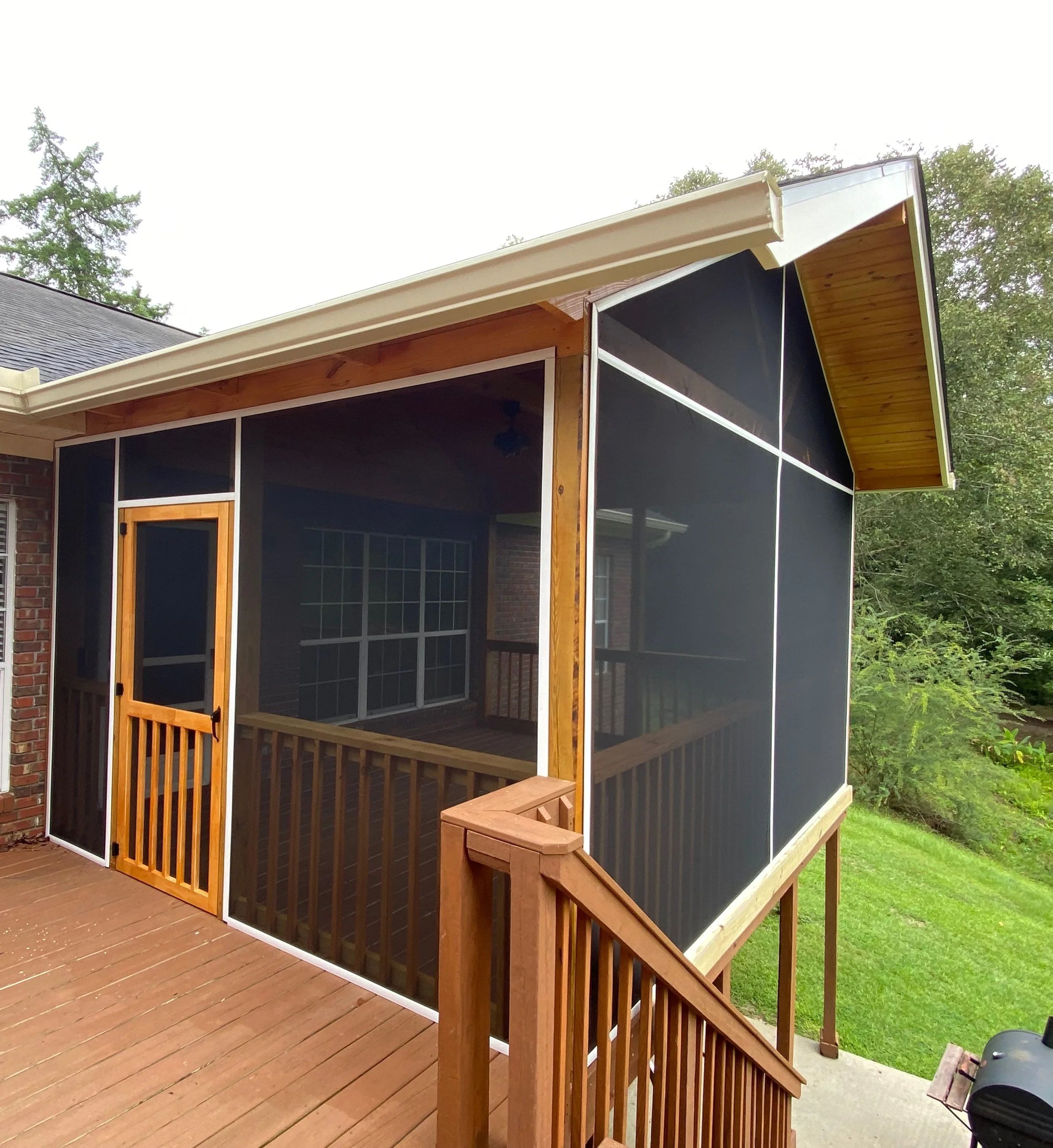A screened-in porch attached to a house with wooden railings and a small door, overlooking a grassy yard and surrounded by trees.