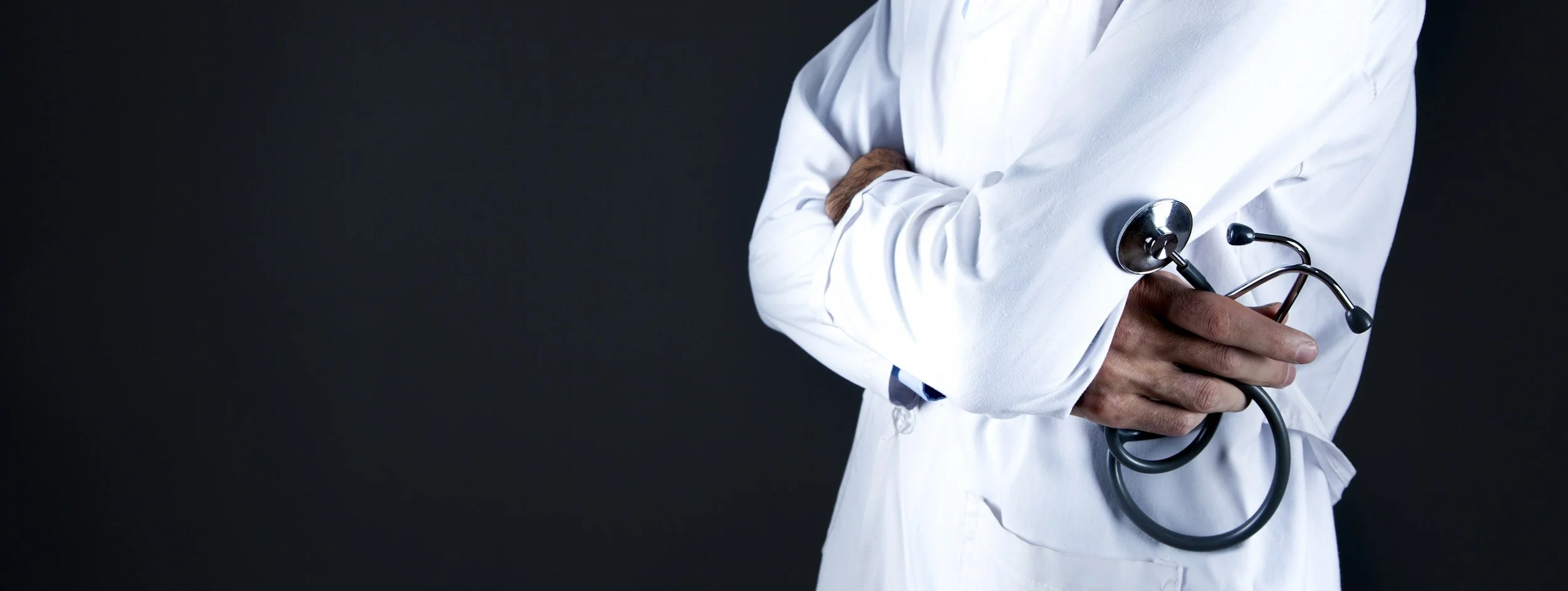 Person in a white medical coat holding a stethoscope in their hand, with arms crossed, against a dark background.