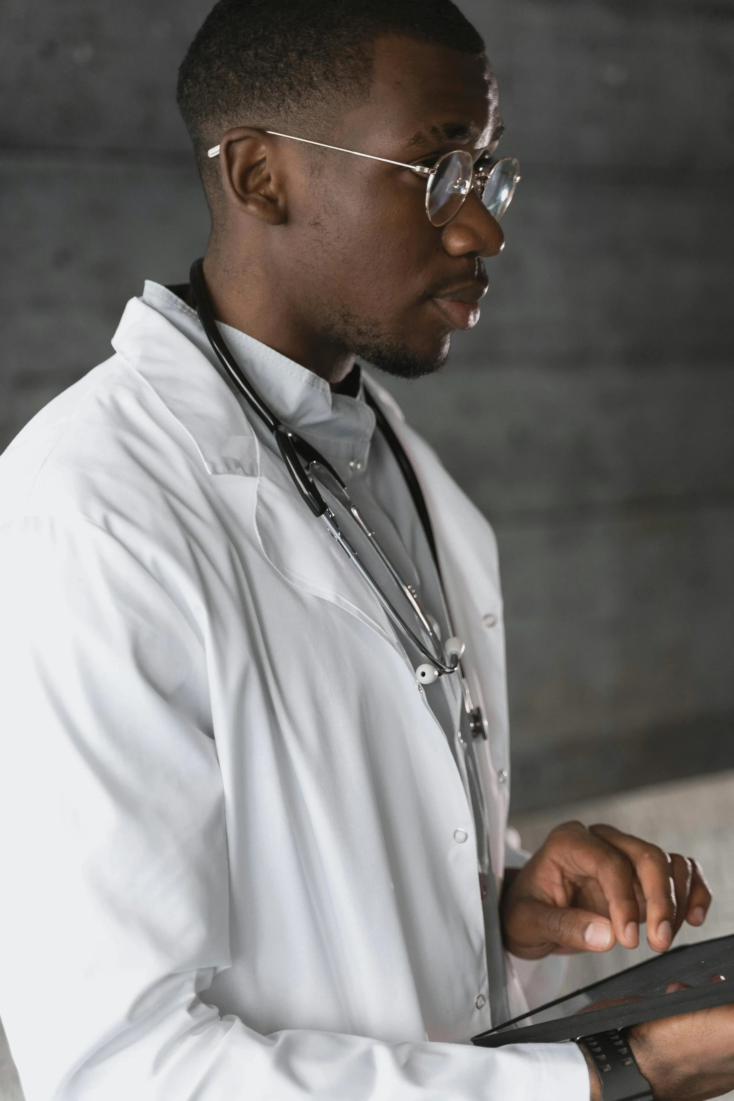 A male healthcare professional wearing glasses and a white coat, with a stethoscope around his neck, using a tablet device.