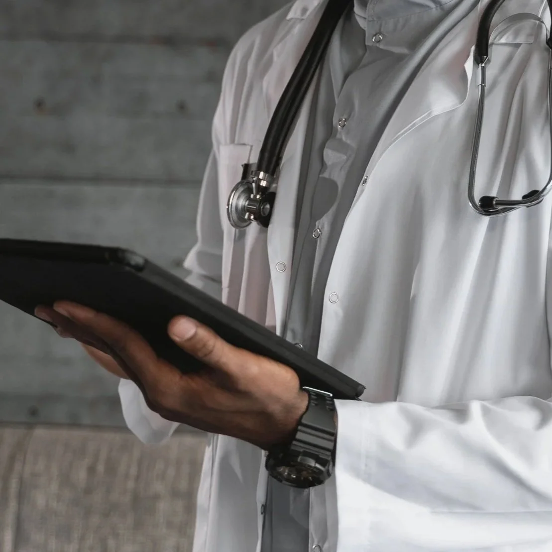 A medical professional wearing a white coat with a stethoscope around their neck, holding a tablet device.