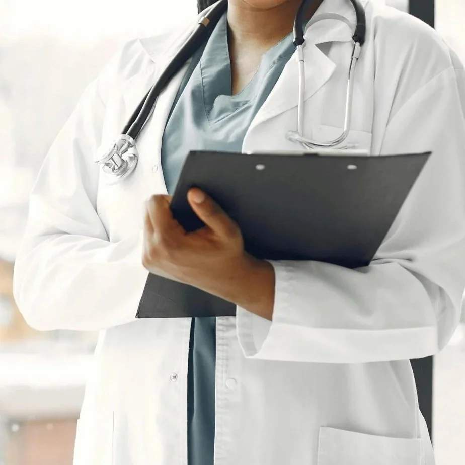 Close-up of a healthcare professional wearing a white lab coat and stethoscope, holding a clipboard in a clinical setting.