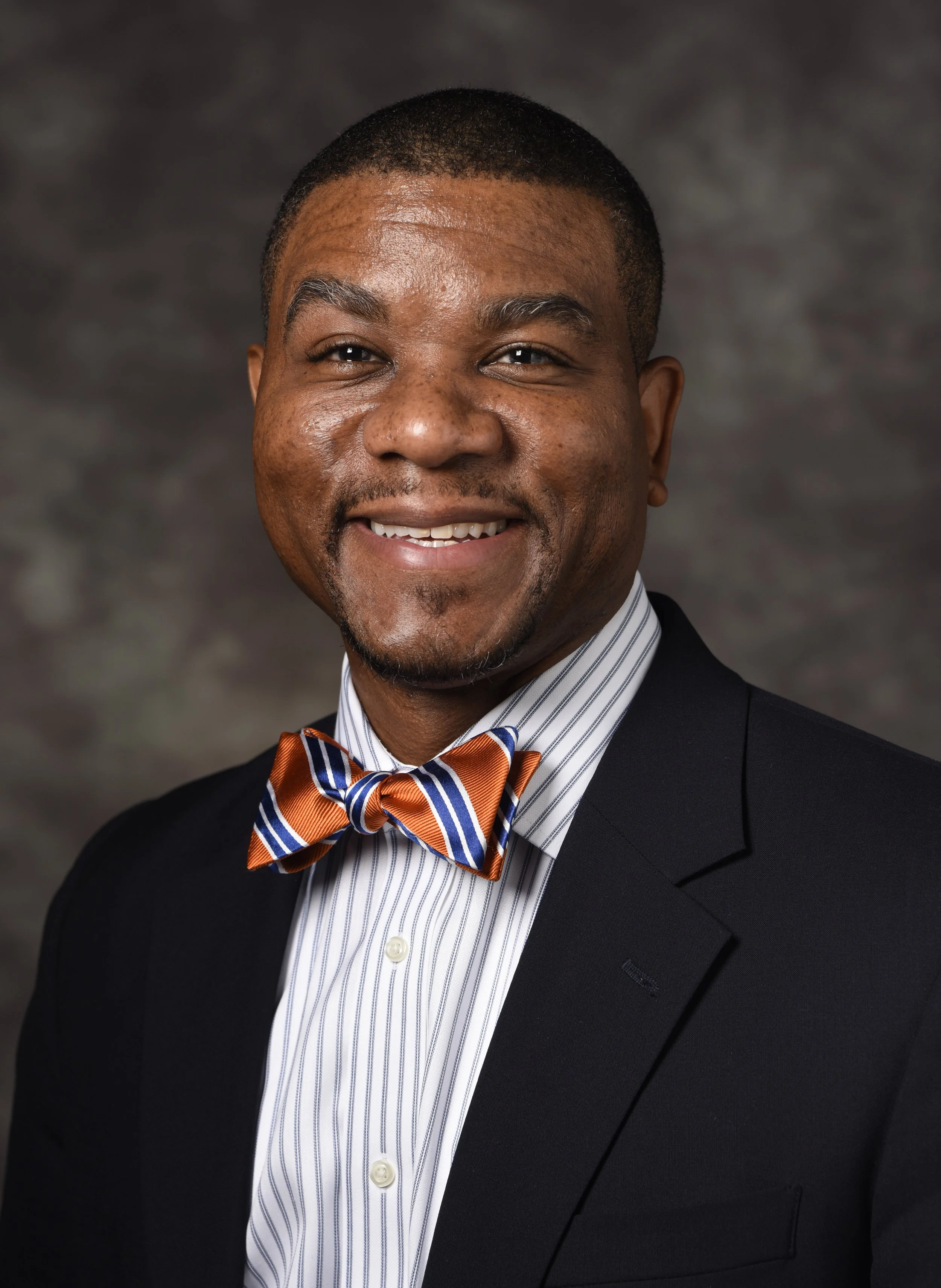 A smiling man in a navy blue suit, white striped shirt, and an orange, blue, and white striped bow tie, standing against a dark, cloudy background.