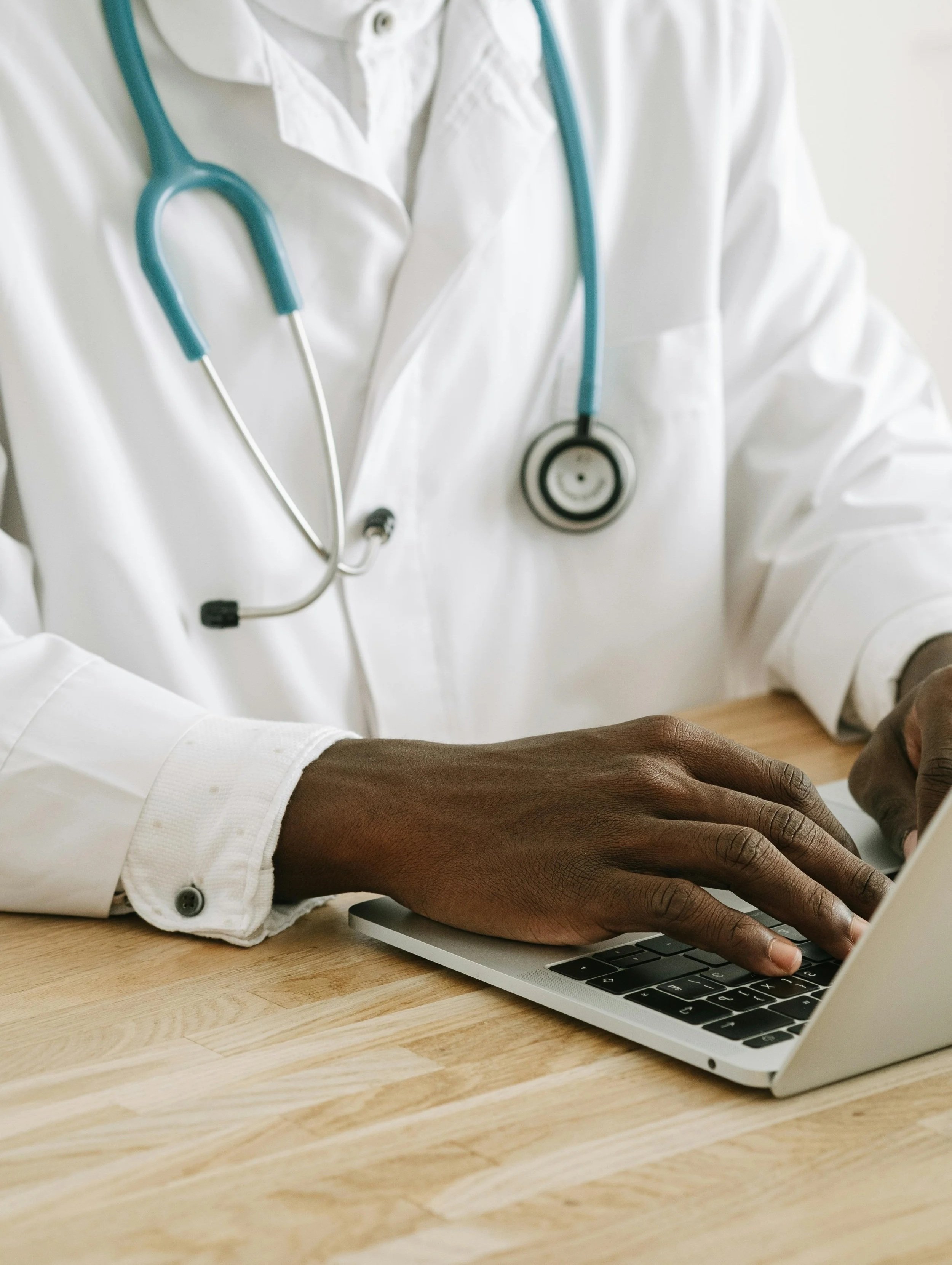 A person with dark skin wearing a white shirt, using a laptop on a wooden table, with a stethoscope hanging around their neck.
