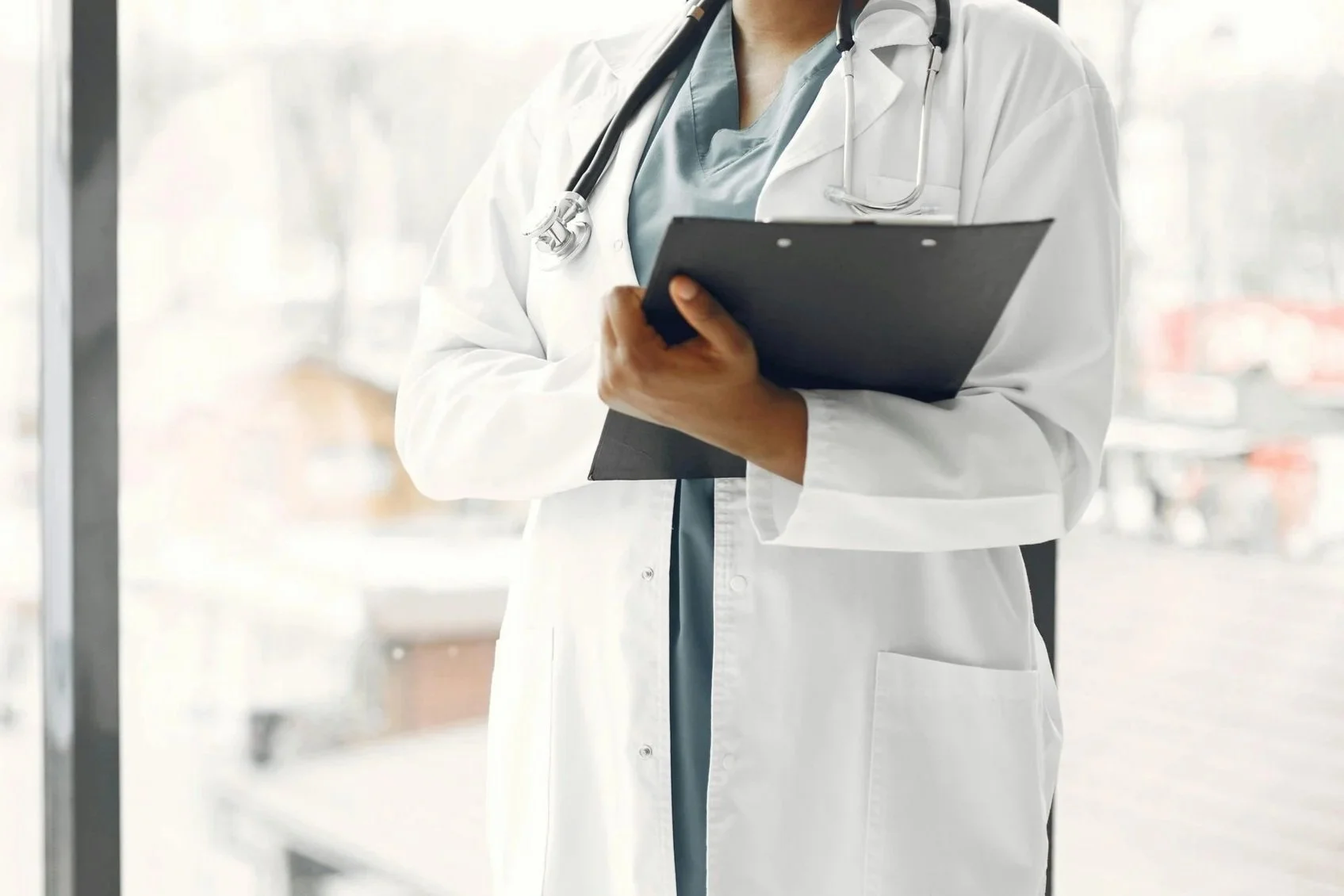 Close-up of a doctor in a white coat holding a clipboard, with a stethoscope around their neck, standing in a bright hospital corridor.