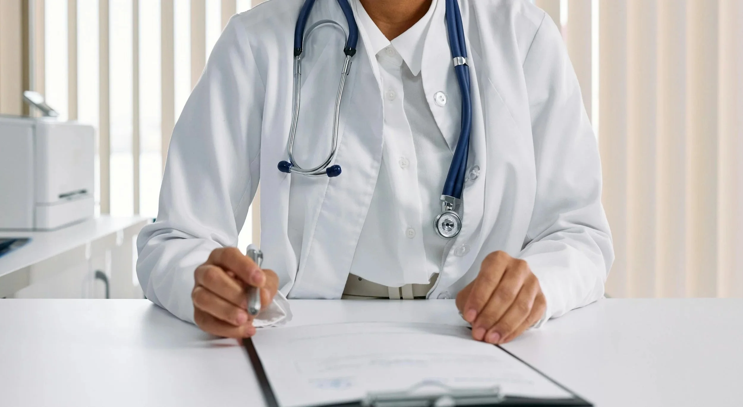 A healthcare professional in a white coat with a stethoscope around their neck sitting at a desk, writing on a notepad in a medical office.