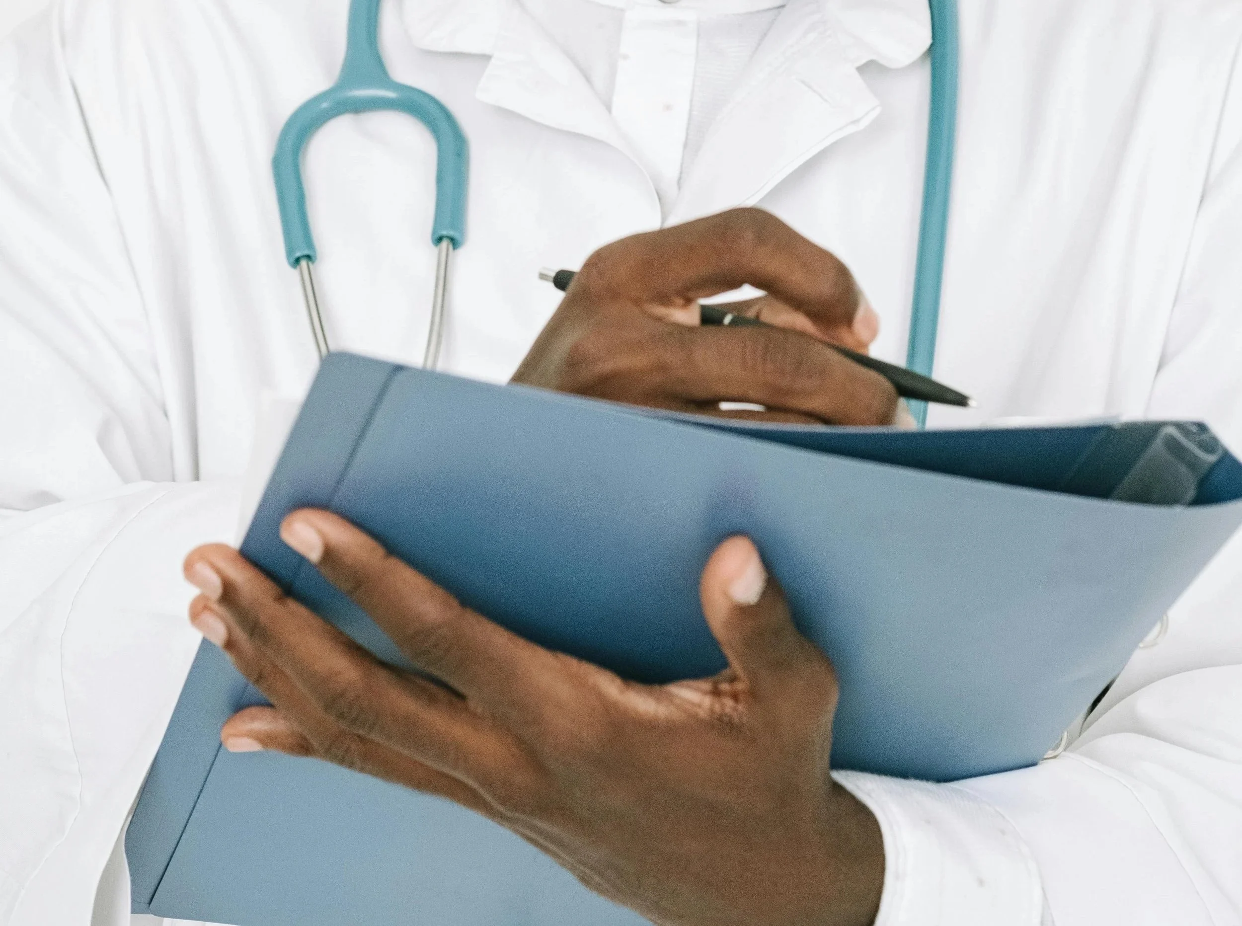 Close-up of a person's hand holding a smartphone over a clipboard. The person is wearing a white lab coat, and a blue stethoscope hangs around their neck.