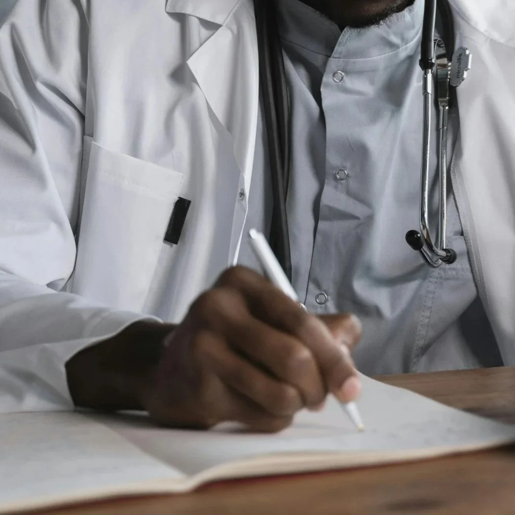 A healthcare professional wearing a white lab coat with a stethoscope around his neck is writing in a notebook with a pen.