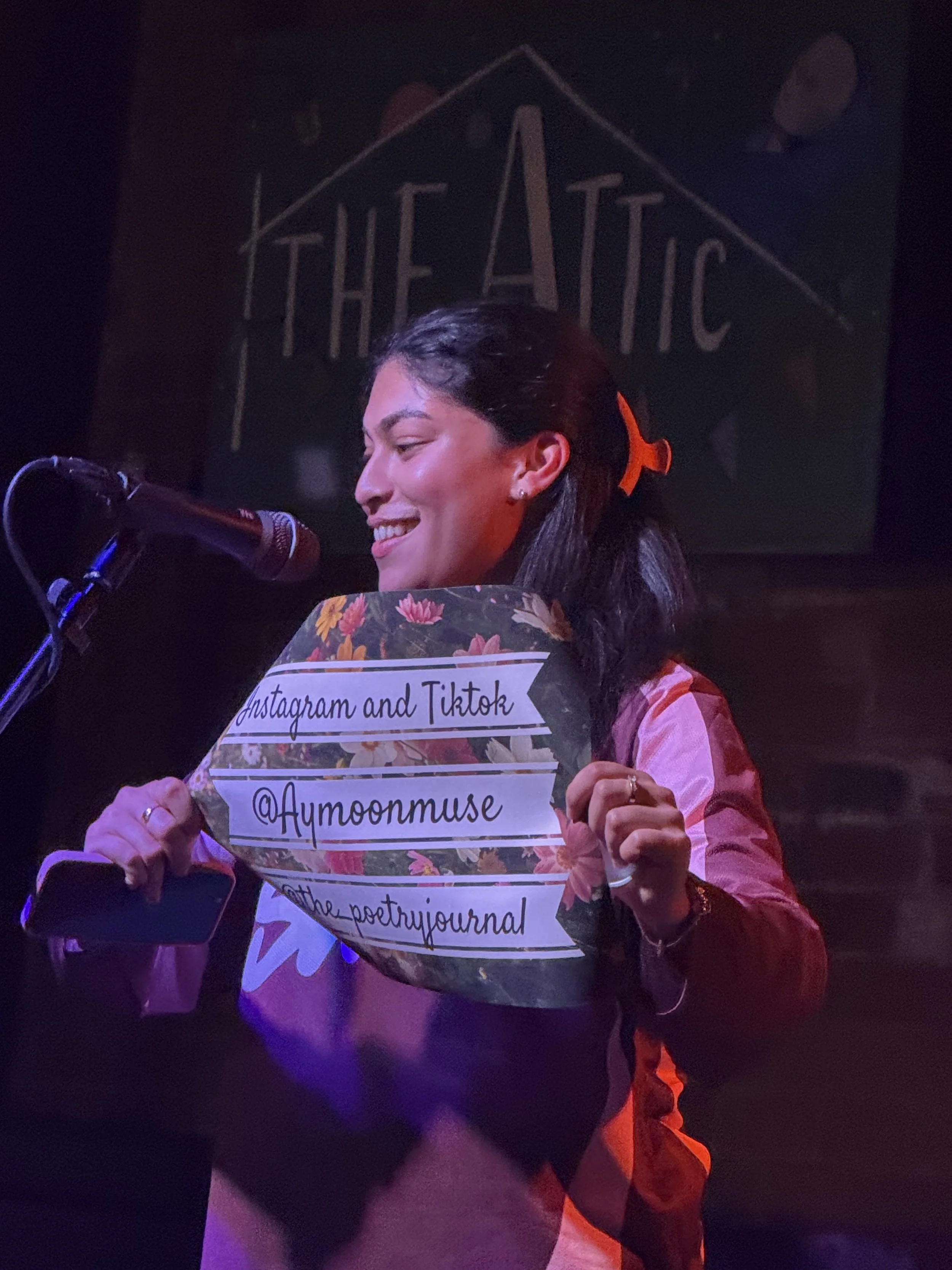 A picture of the founder of Samajh, holding up a poster at an open mic with the handles to her instagram on it, smiling, standing at the mic