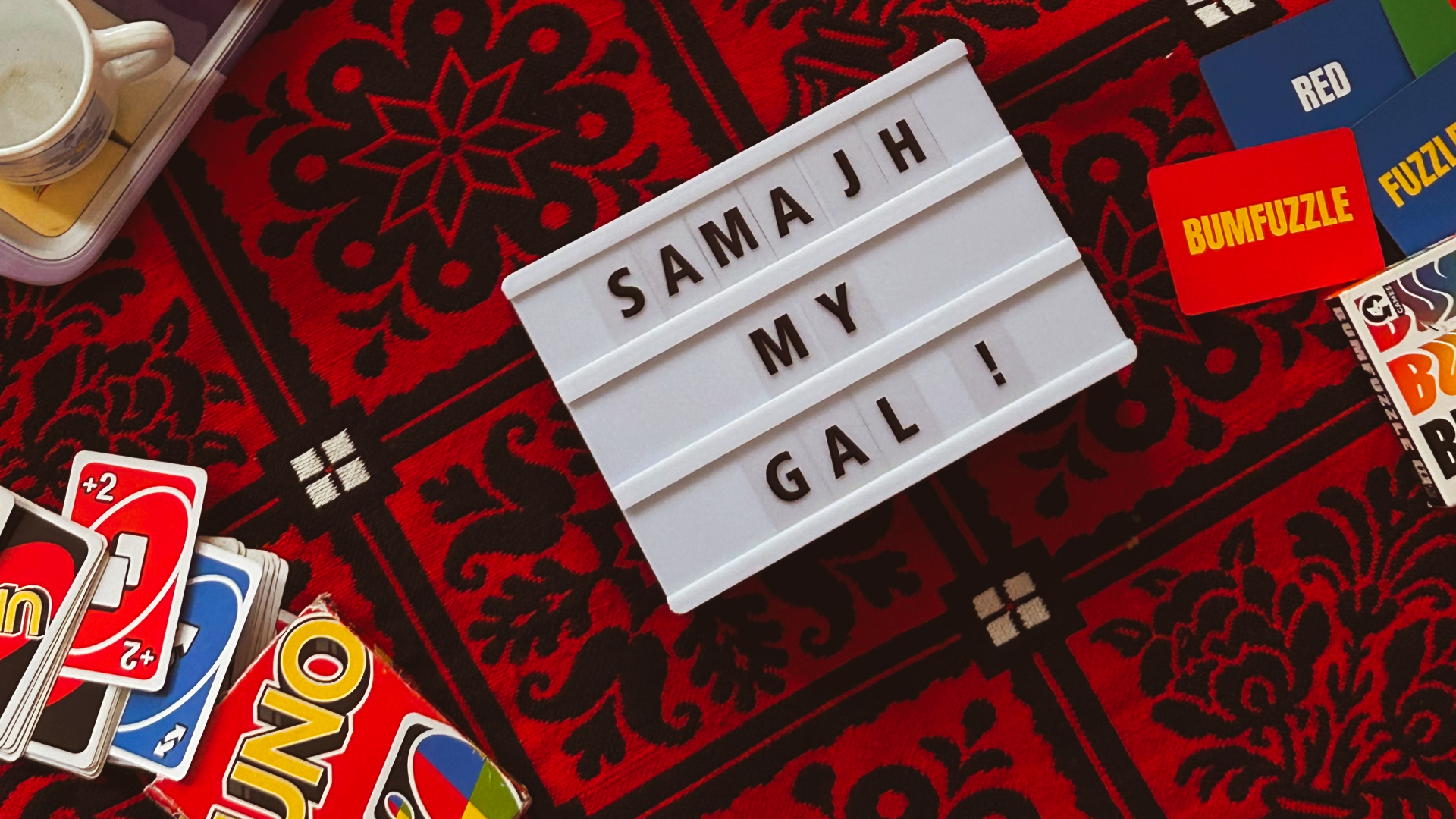 A letter and lightbox with the words 'Samajh My Gal', on a Persian/ Pakistani rug, surrounded by games like UNO and Bumfuzzle and a tray of clay pots