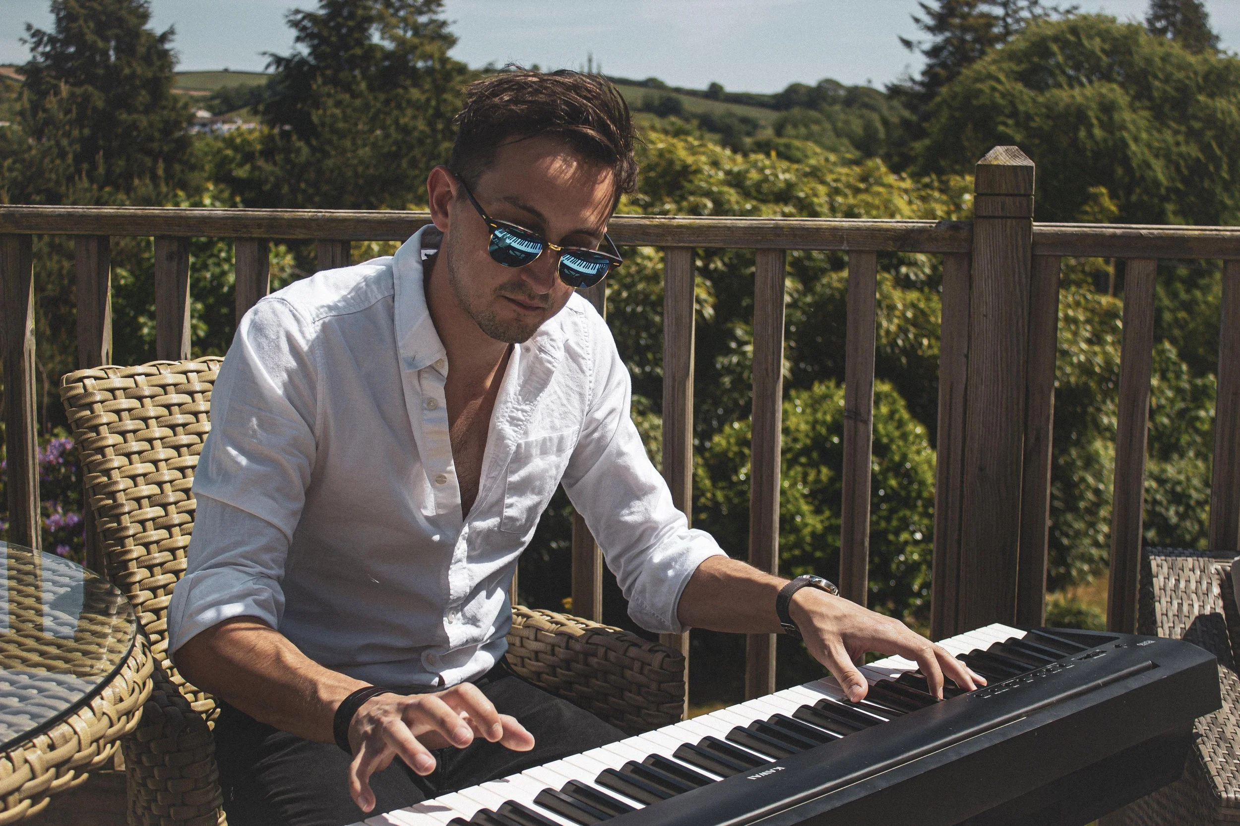 A man wearing sunglasses and a white shirt playing a keyboard on an outdoor patio with a wooden railing, trees, and blue sky in the background.
