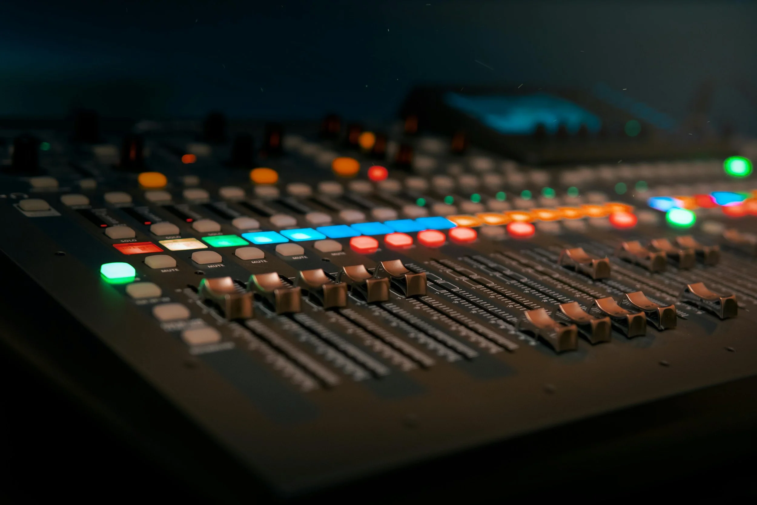 A professional audio mixing console with colorful illuminated buttons, sliders, and knobs in a dark environment.