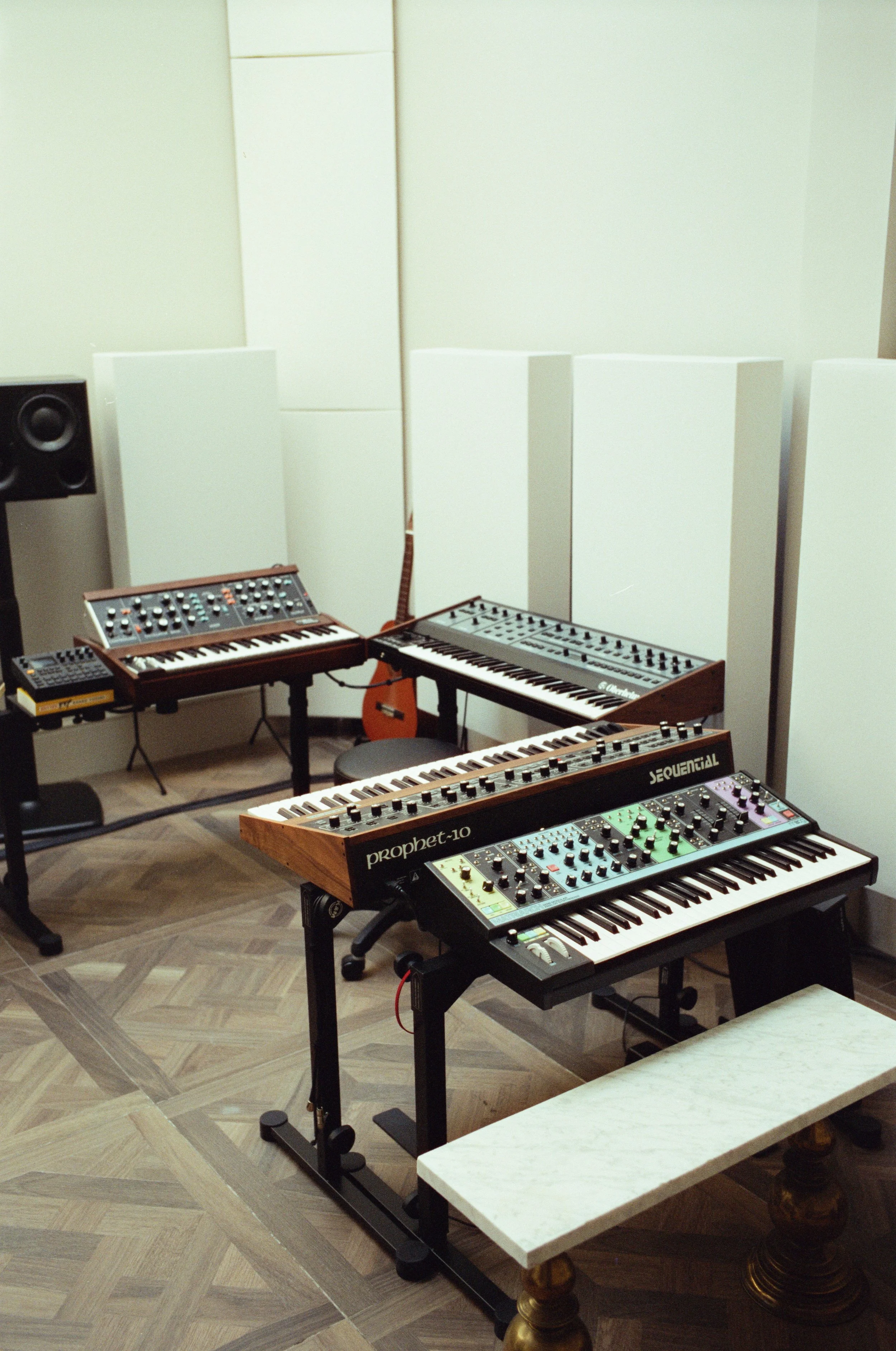 A music studio with multiple synthesizers, speakers, and a guitar. The room has wooden flooring and white walls with soundproofing panels.