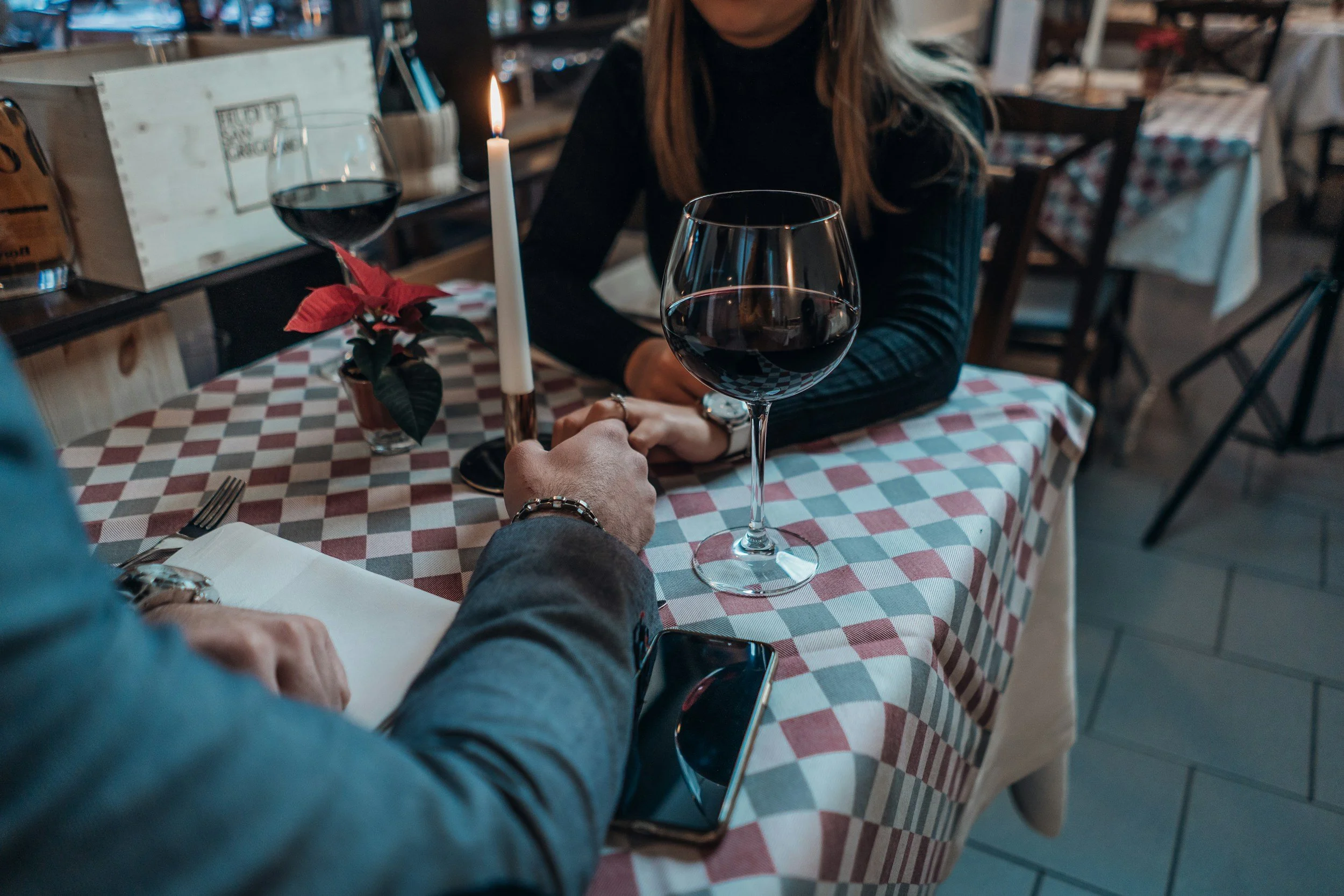 A romantic dinner setting with a person holding a lit candle and a woman sitting across the table. The table has a checkered tablecloth, a glass of red wine, a potted poinsettia plant, and a notebook. The background shows other tables and chairs in a cozy restaurant.