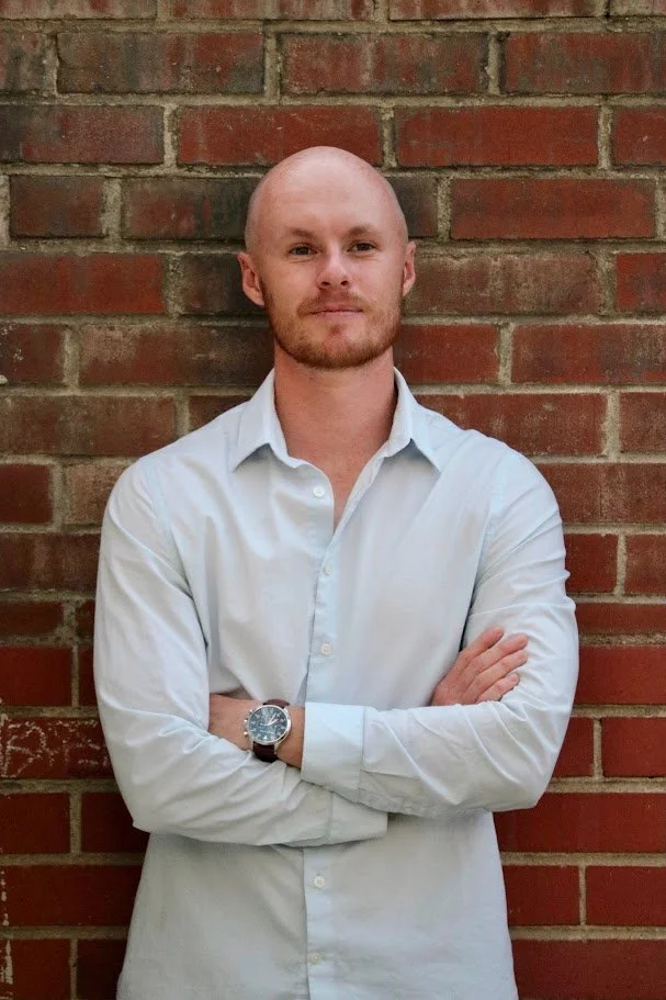 A man with a shaved head and beard, wearing a white button-up shirt and a wristwatch, stands with arms crossed in front of a red brick wall.