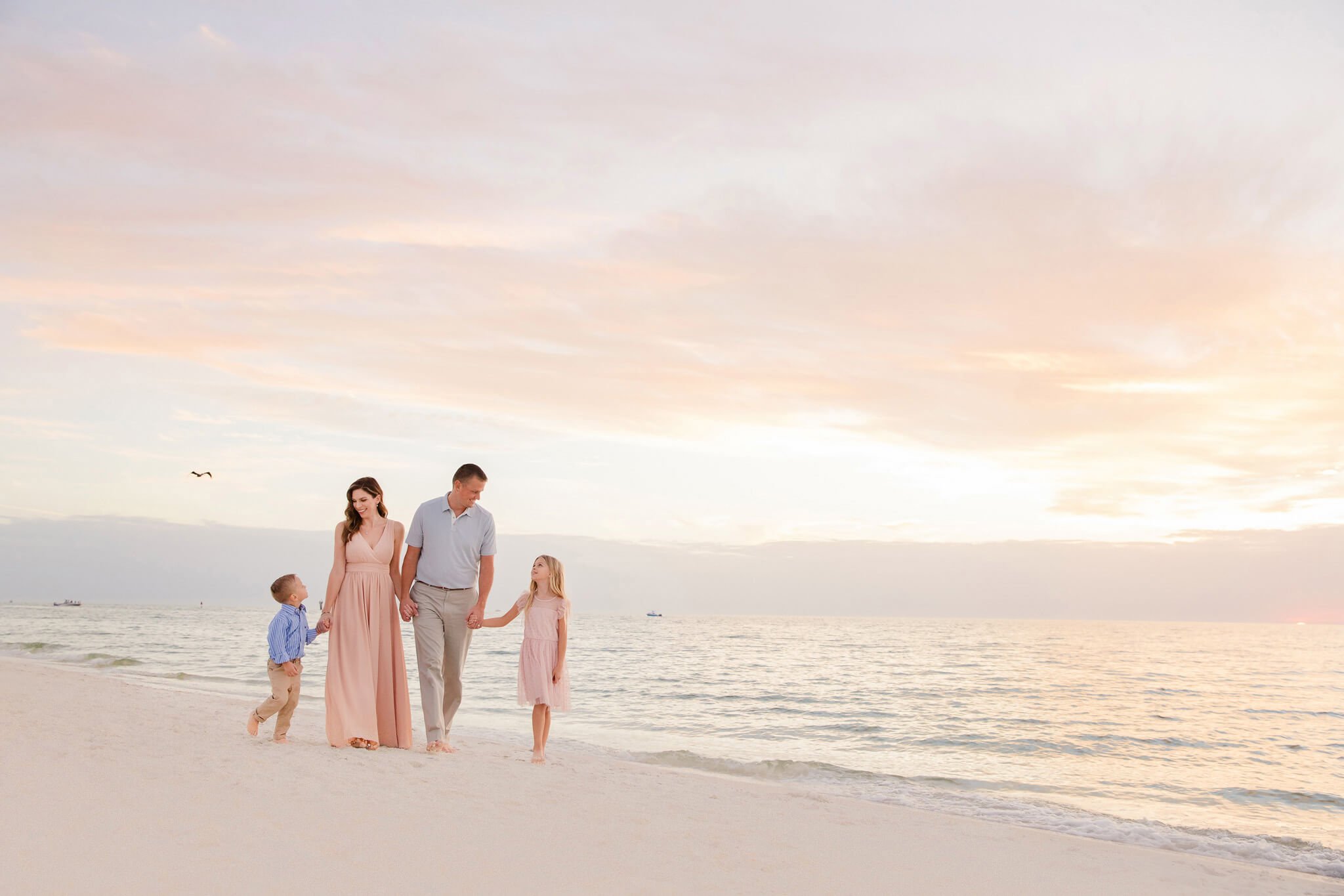 A family of four walking on a beach at sunset, holding hands, with calm ocean waves and a pastel-colored sky in the background.