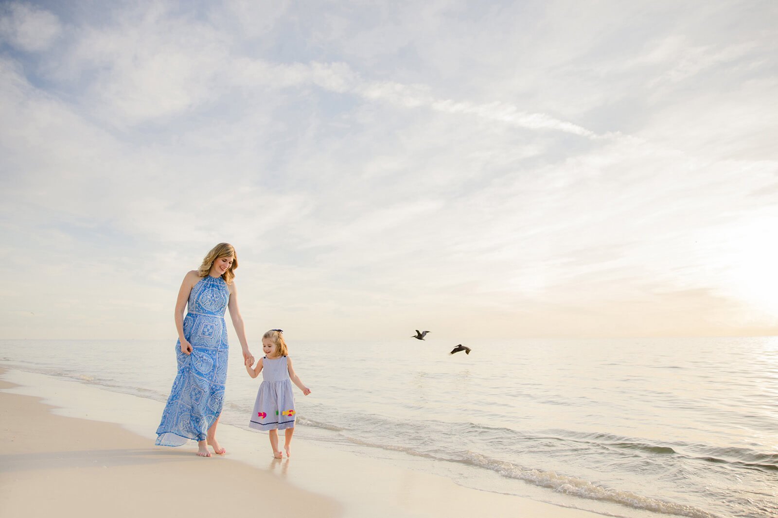A woman and a young girl walking hand in hand along the beach near the water, with a cloudy sky and a few birds flying overhead during a Naples FL beach photography session.
