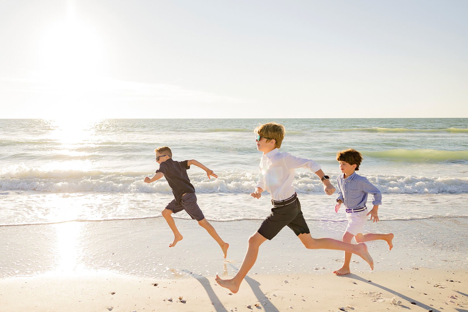 Three boys running on the beach near the ocean, with waves and the sun in the background during family beach portraits Naples FL.
