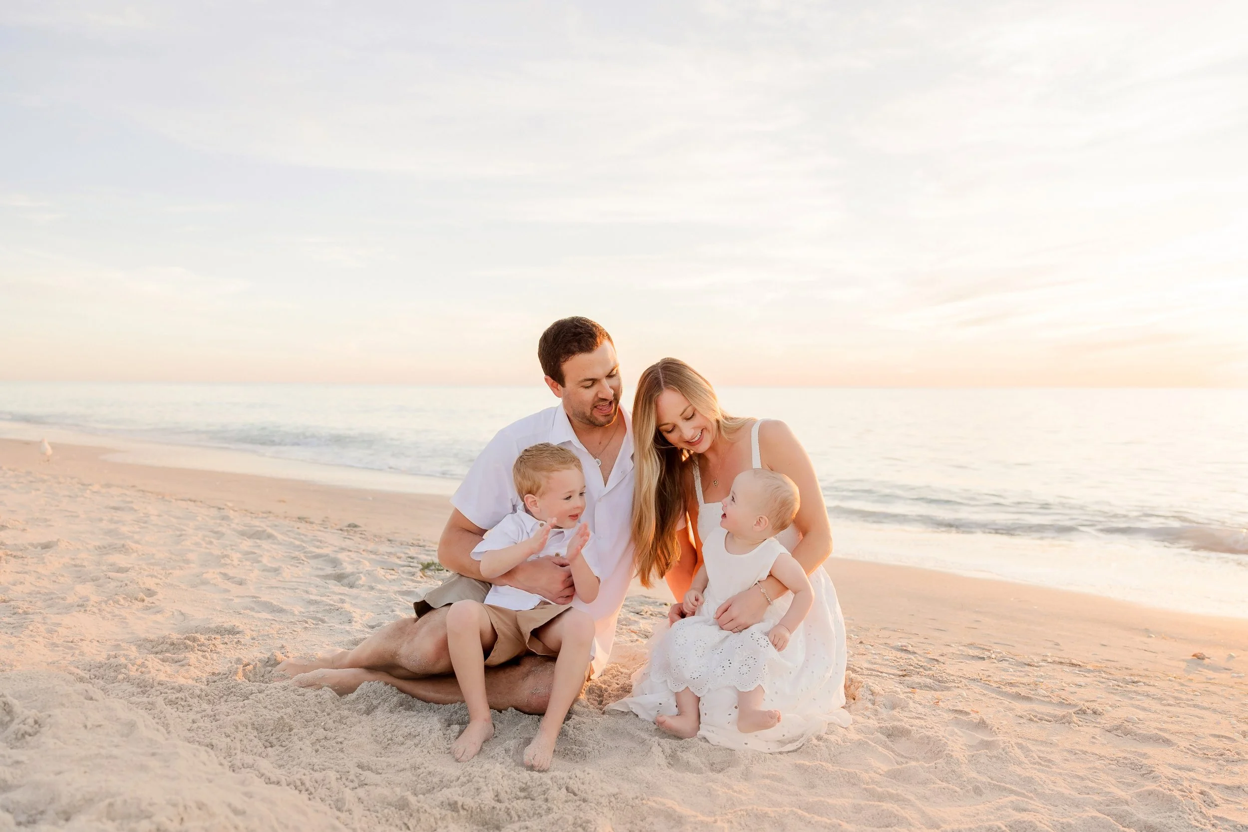 A family of four enjoying time on the beach at sunset, sitting on the sand and smiling at each other taken by a Naples photographer.
