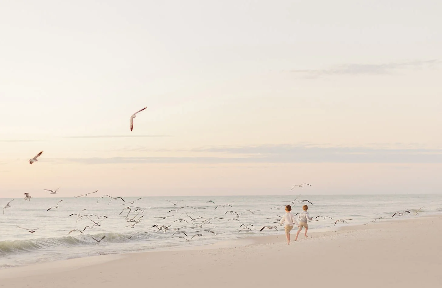 Two children holding hands and running along the beach near the shoreline as seagulls fly overhead during sunset.