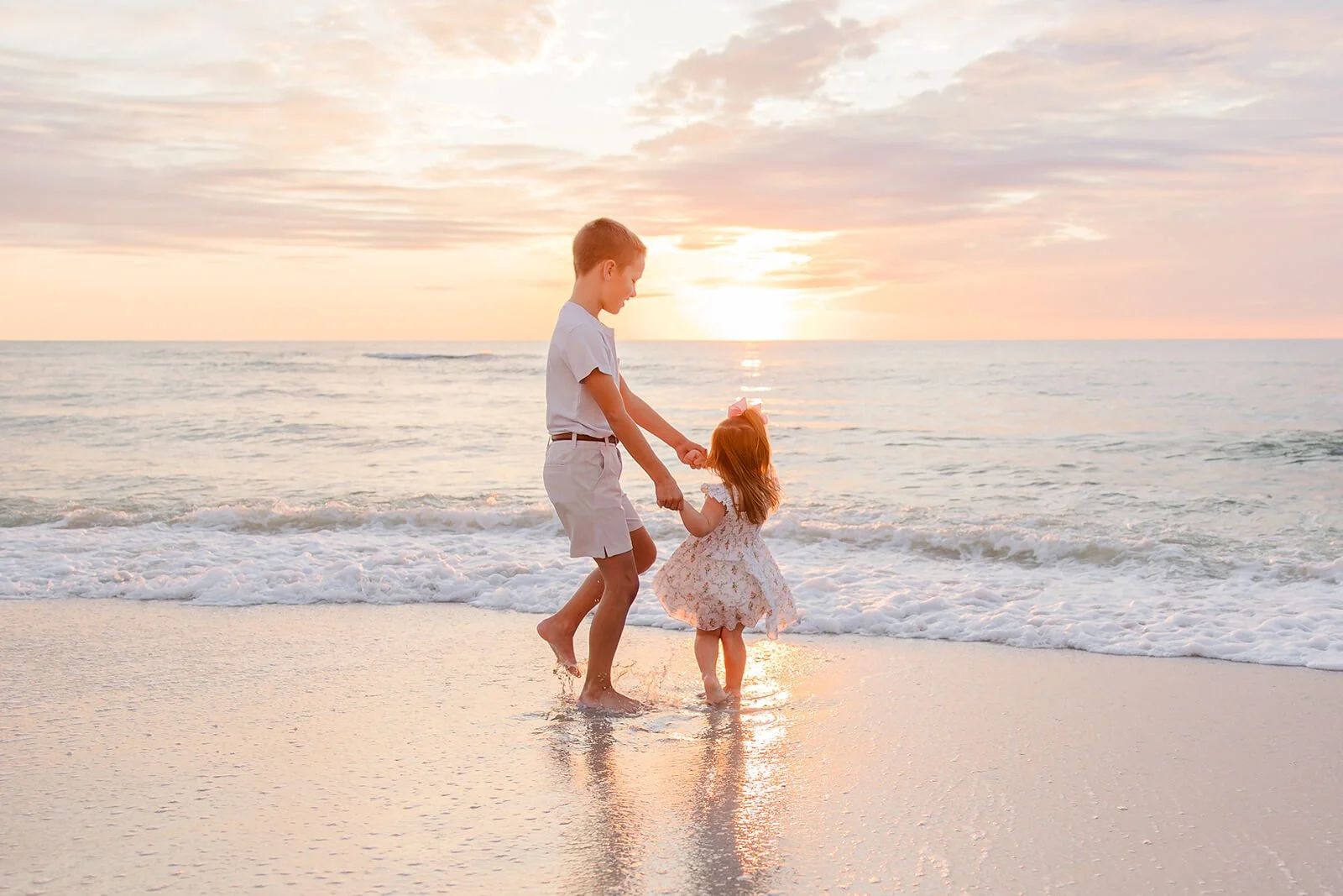 A boy and a girl playing together on the beach during sunset, holding hands, with waves at their feet for a Naples beach family portrait.
