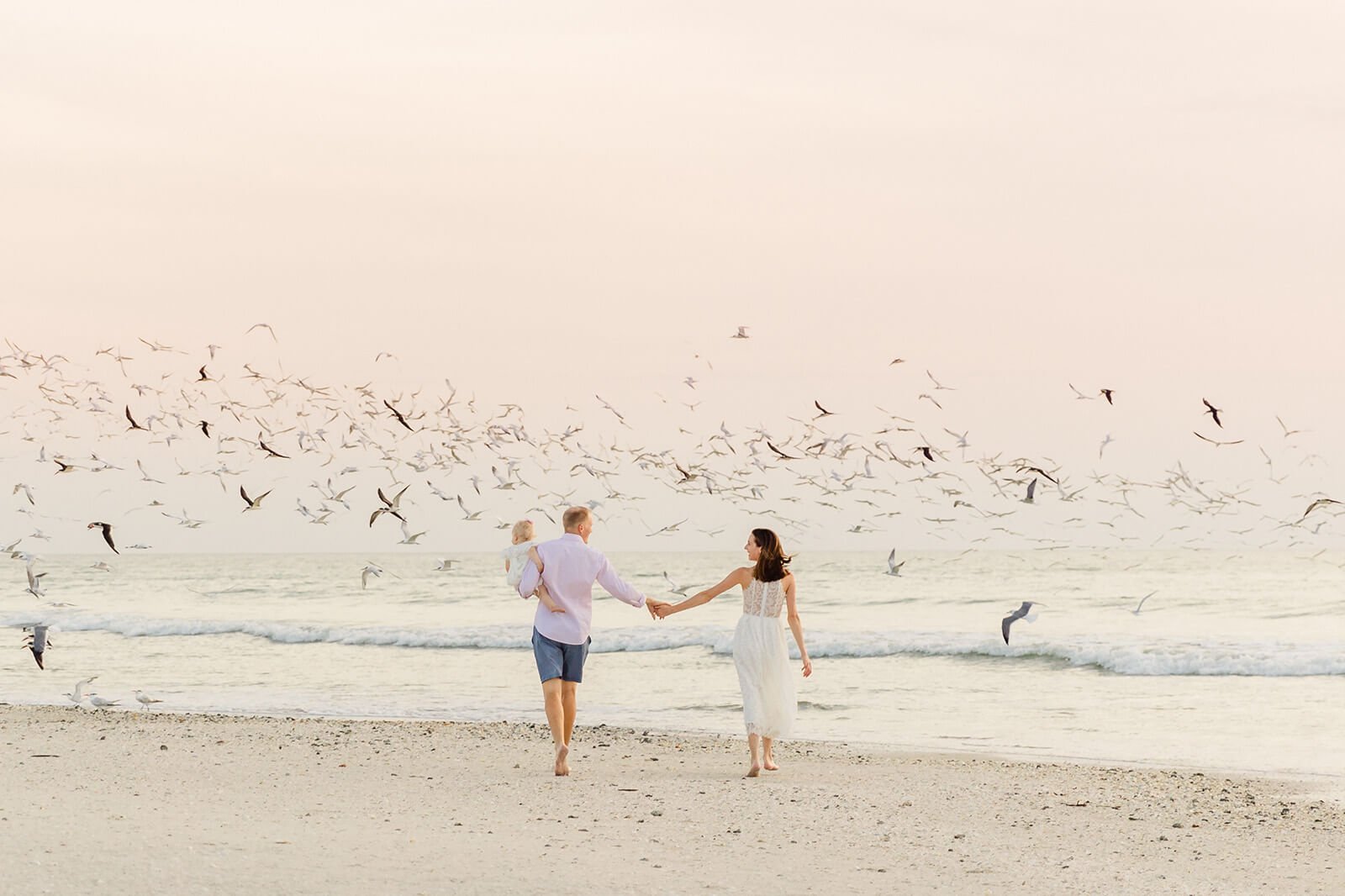 A family walking on a beach with seagulls flying overhead during sunset during a Naples beach portrait session.