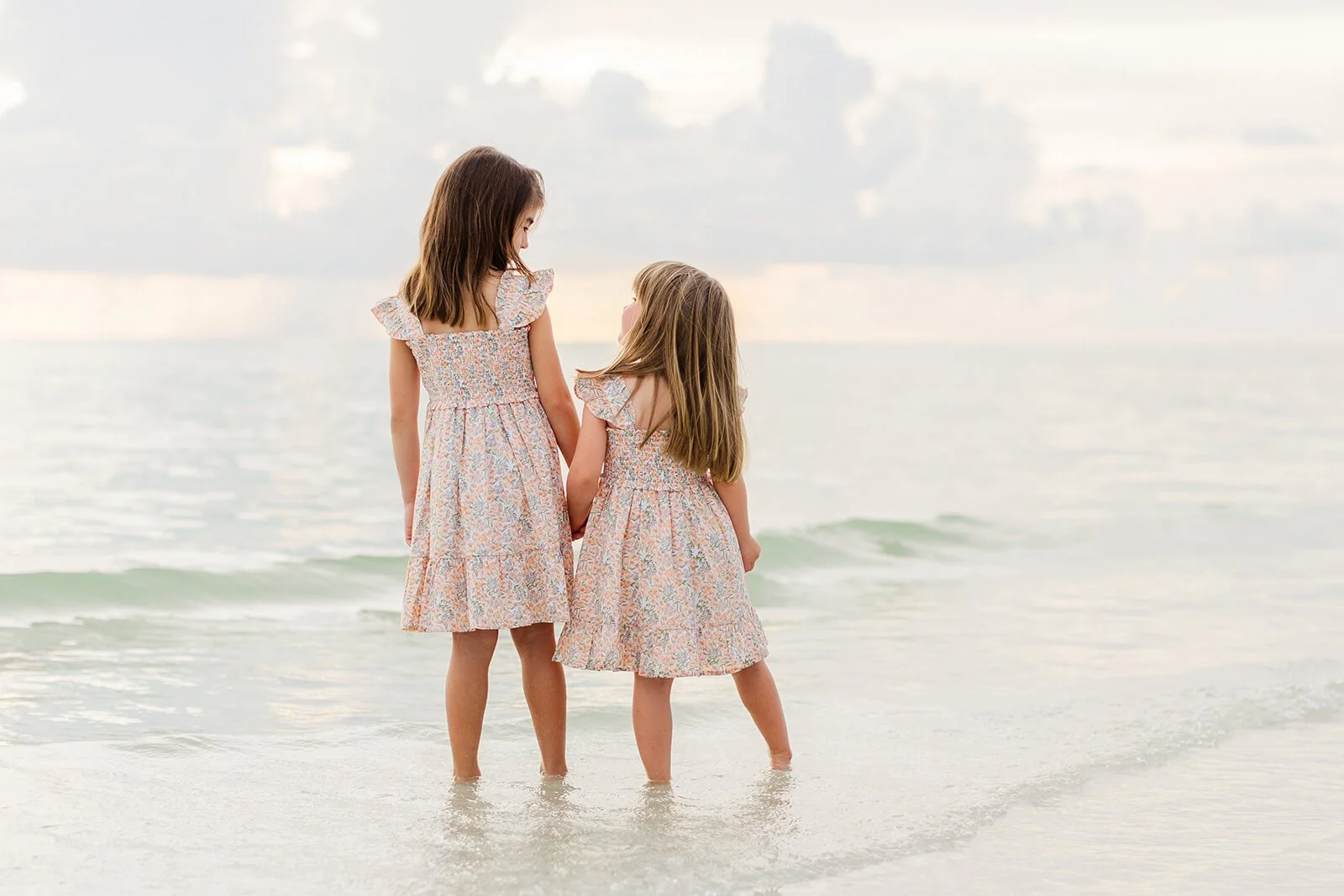 Two young girls in matching floral dresses standing in shallow water at the beach, holding hands and looking at each other, with a cloudy sky in the background photographed on a Naples Florida beach.