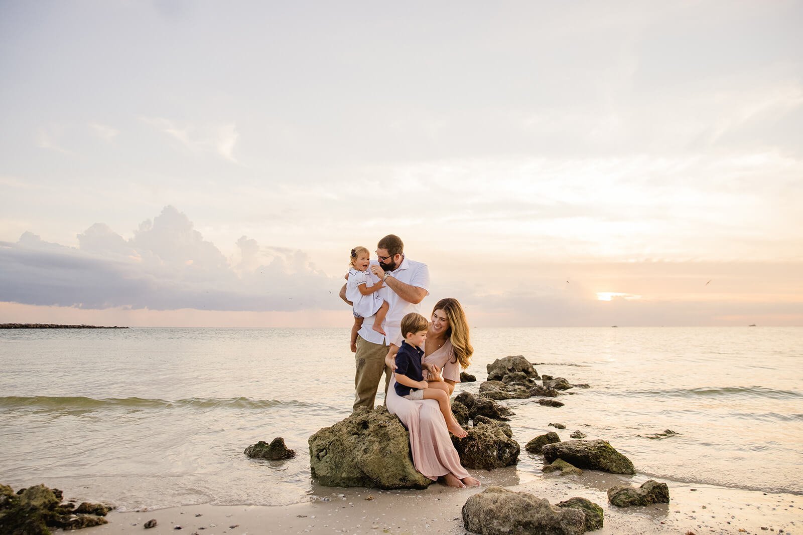 A family of four on a rocky beach at sunset, with the ocean and cloudy sky in the background on the Gulf coast by a photographer in Naples FL.