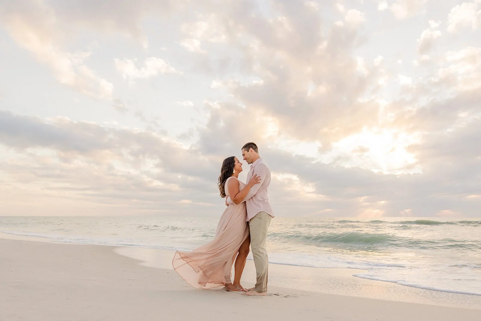 A couple in formal attire standing on the beach at sunset, gazing into each other's eyes, with the ocean and cloudy sky in the background during a Naples Fl engagement session.