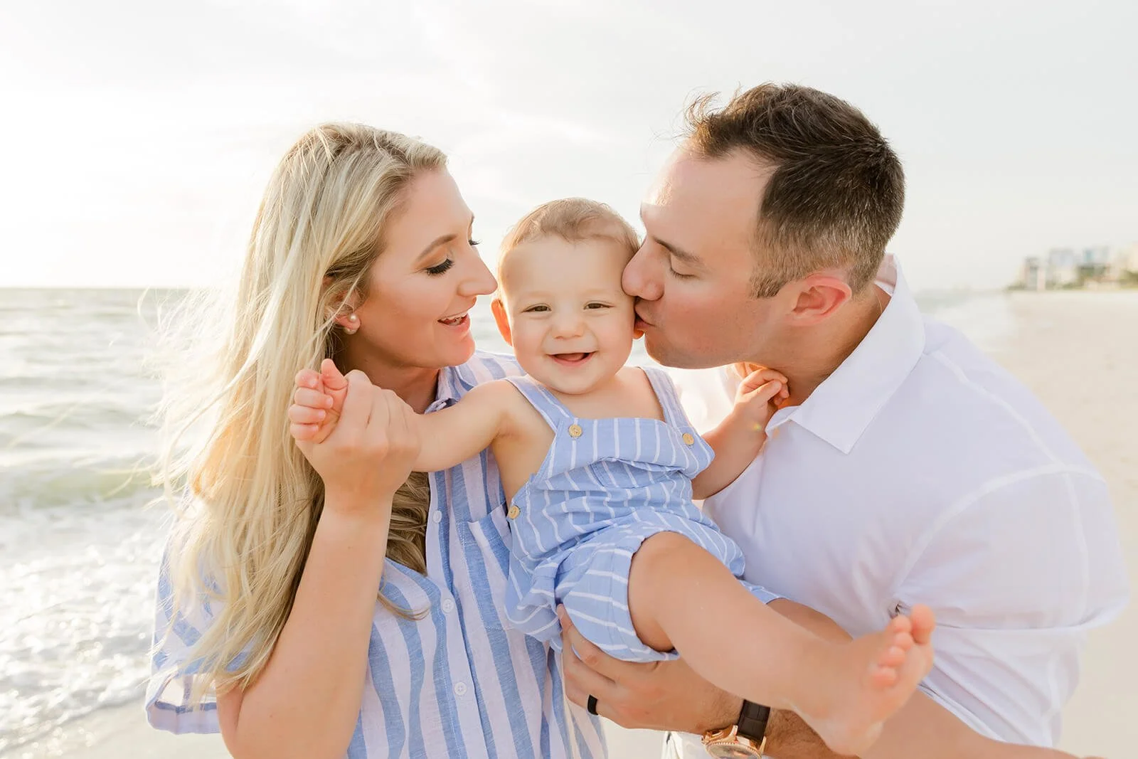 A happy mother, father, and a baby, enjoying time together on the beach with ocean waves in the background. The baby is smiling while being held by the parents, and the father is giving the baby a kiss on the cheek while taking Naples beach photos.
