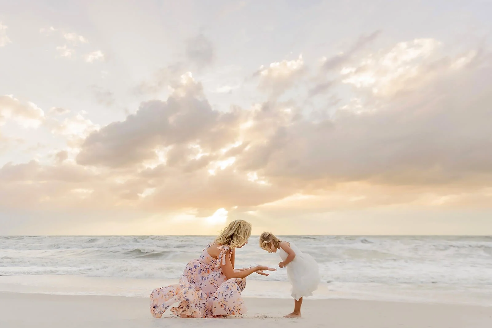A woman and a young girl on the beach, with the woman kneeling and both looking at something in her hand, during sunset with clouds in the sky during a Naples beach family portrait session.